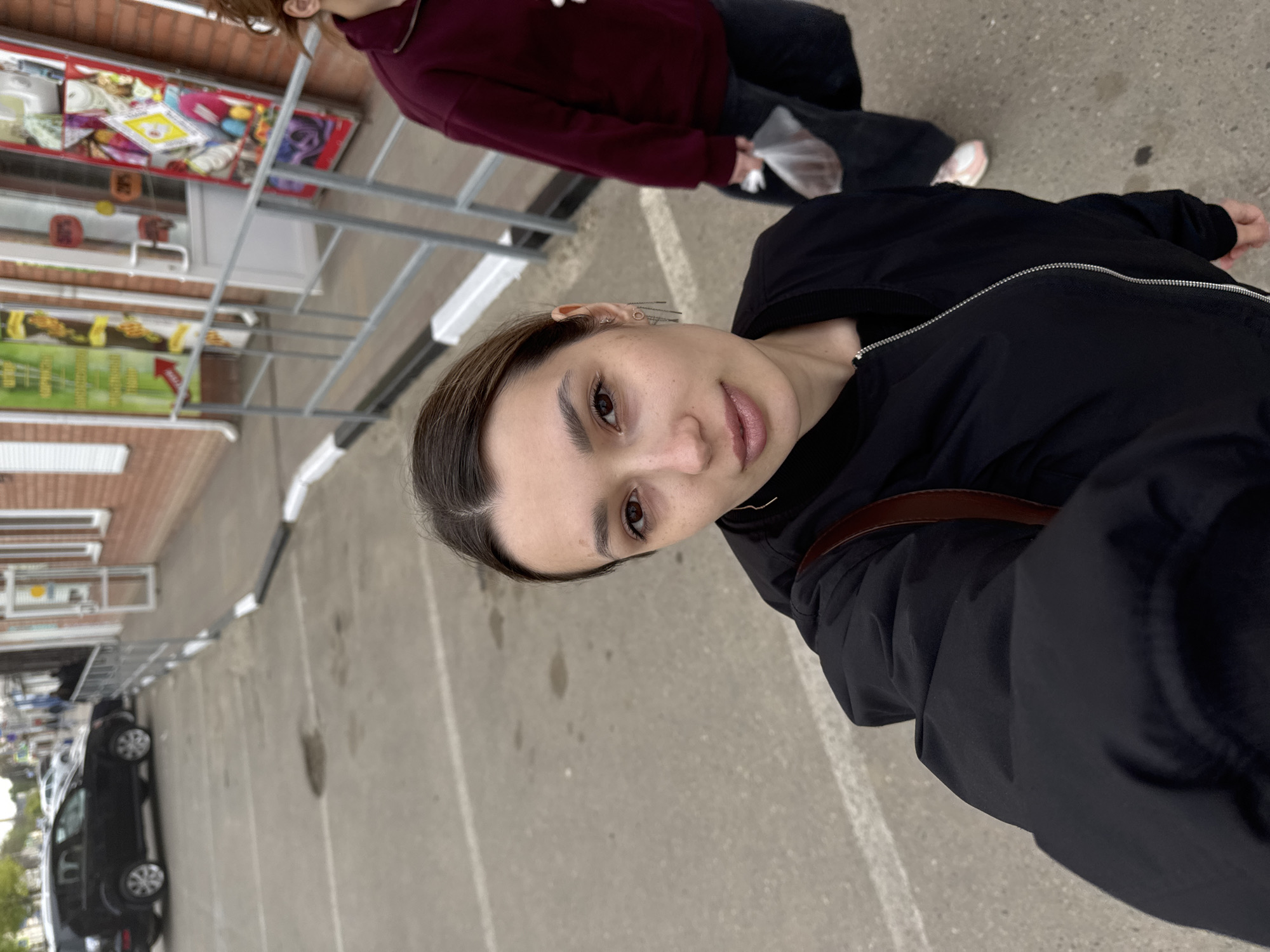 A young woman is featured prominently in the foreground of this outdoor selfie, angled from her right side looking towards the camera. She has dark hair pulled back, light brown eyes, and a neutral to slightly pleasant expression. She is wearing a black zippered jacket, with a brown strap, possibly from a bag, visible over her shoulder. Small hoop and stud earrings are visible in her left ear.
In the mid-ground, another person is partially visible, walking away from a row of shops. This individual is wearing a burgundy long-sleeved top, dark pants, and is holding a white plastic bag.
The background reveals an outdoor commercial setting, likely a paved area such as a parking lot or a wide sidewalk. To the left, a series of brick-colored buildings house various small shops or stalls, characterized by display windows showcasing products. A metal railing structure runs along the front of these buildings. Further in the background, a black car is parked. The ground is concrete with visible white painted lines.
The lighting suggests it is daytime, with soft, diffused light, indicating an overcast sky or an area with indirect sun. Some indistinct text and graphics are visible on the shop signs in the background, including orange price tags that vaguely show numbers like "16" and "35," and a yellow sign with a graphic.