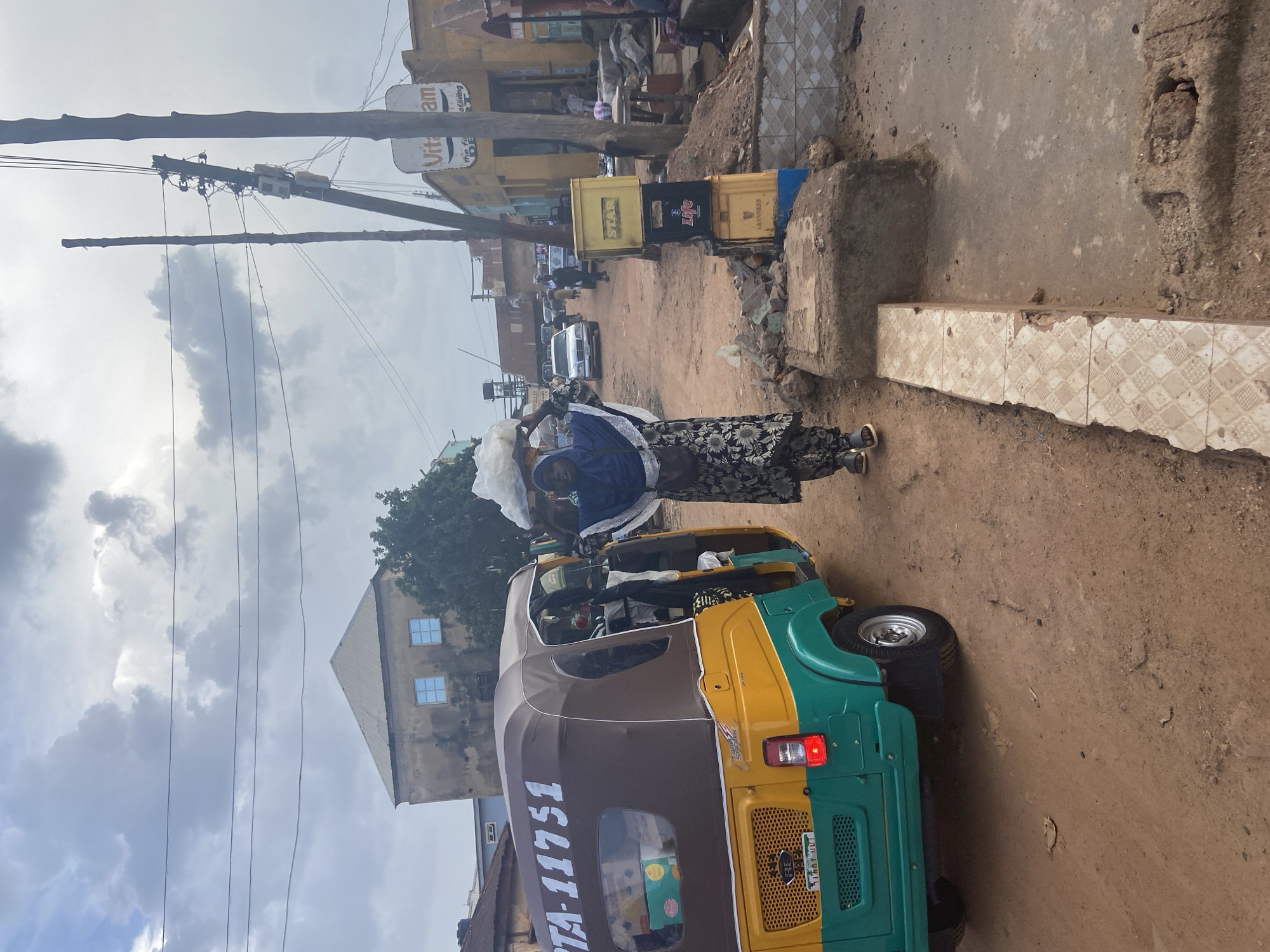 In Bukuru, Nigeria, a woman carries a large load of goods on her head while walking down a dirt road. She is wearing a blue headscarf and a dark floral pattern dress. To her left, a yellow and green auto-rickshaw, with the license plate "KTA-11751," is parked. The rickshaw has a brown canvas cover and is packed with various items. On the right side of the road, a stack of yellow crates, some labeled "STAR" and "GUINNESS," are piled high. A storefront with a sign reading "Vita am" is visible in the background, along with other buildings and power lines. The sky is overcast with clouds.