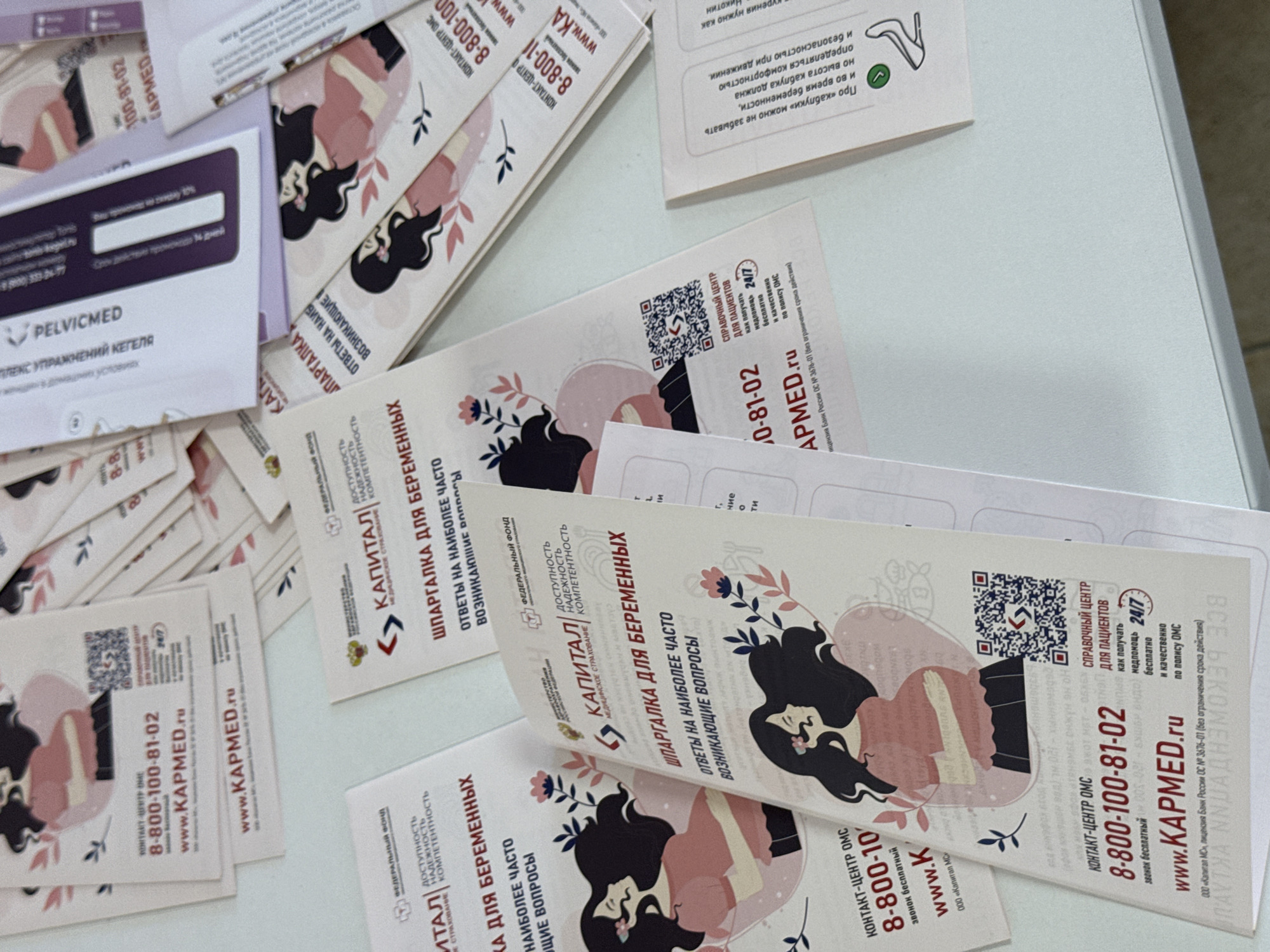 The image shows a top-down view of a collection of printed materials, likely brochures or flyers, scattered across a light-colored surface. The materials feature illustrations of women, some appearing pregnant or cradling a baby, in a soft, pastel color palette.
The dominant text visible is in Russian. Many of the flyers contain contact information, including phone numbers like "8-800-100-81-02" and website addresses such as "www.KAPMED.ru". Other text includes phrases like "ШПАРГАЛКА ДЛЯ БЕРЕМЕННЫХ" (Cheat sheet for pregnant women), "ОТВЕТЫ НА НАИБОЛЕЕ ЧАСТО ВОЗНИКАЮЩИЕ ВОПРОСЫ" (Answers to the most frequently asked questions), and "СПРАВОЧНЫЙ ЦЕНТР ДЛЯ ПАЦИЕНТОВ" (Information center for patients). Some flyers also mention "КАПИТАЛ" and "МЕДИЦИНСКОЕ СТРАХОВАНИЕ" (Medical insurance), suggesting a connection to healthcare services. One flyer depicts a graphic of a high-heeled shoe with a Russian caption about "heels" and "pregnancy."
There is also a product name visible: "PELVICMED" with the text "КОМПЛЕКС УПРАЖНЕНИЙ КЕГЕЛЯ В ДОМАШНИХ УСЛОВИЯХ" (Kegel exercise complex at home). QR codes are present on several of the flyers.
The overall impression is of informational materials related to pregnancy, women's health, and potentially medical services available in Oktyabrskoye, Russia. The arrangement of the papers suggests they have been recently handled or distributed.