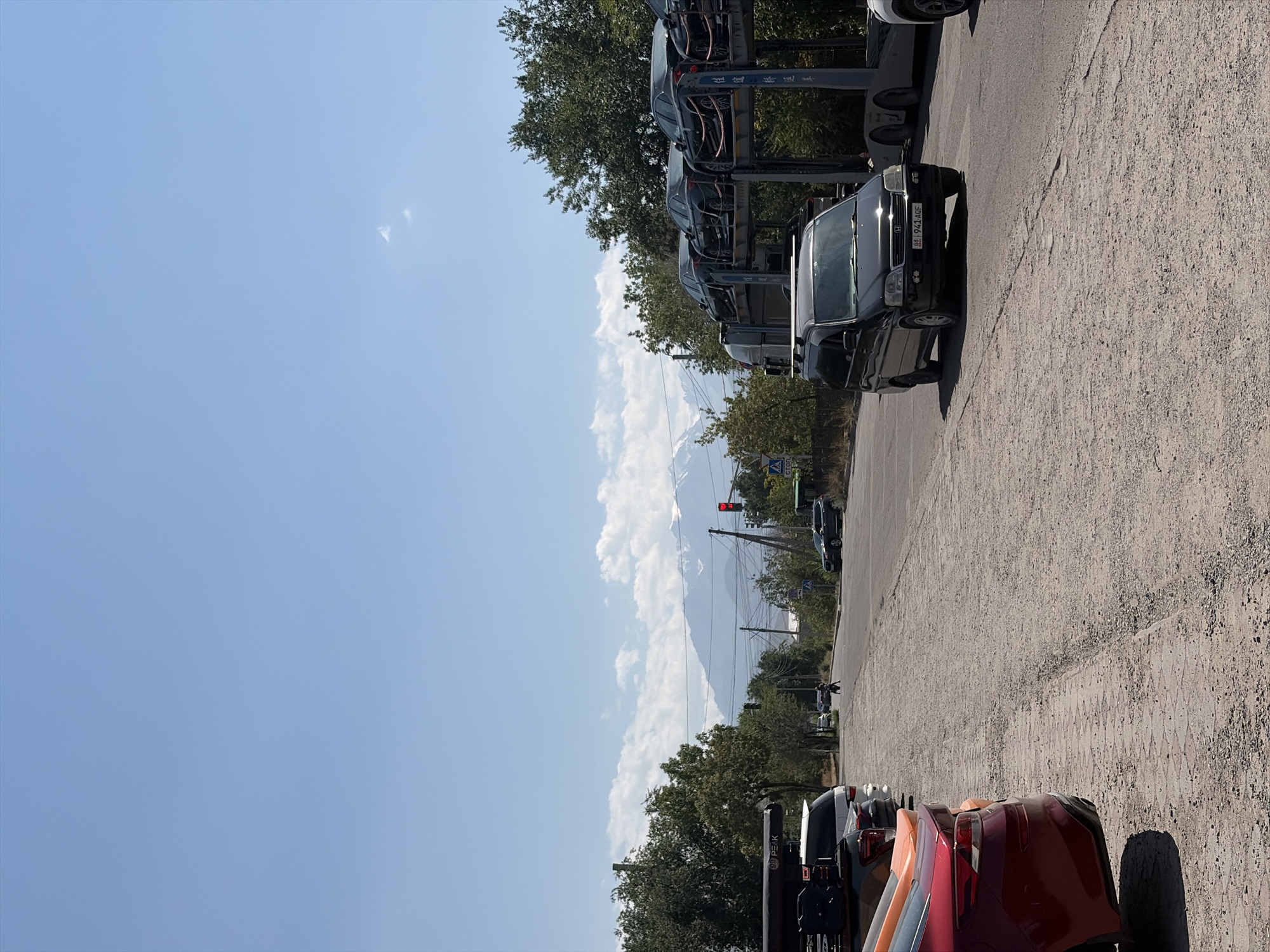 The image depicts a street scene in Bishkek City, Kyrgyzstan, on a clear day with a bright blue sky and scattered white clouds. In the foreground, a dirt and gravel road is visible, with several parked cars lining the left side. These cars include a red hatchback, an orange vehicle, a white SUV with a roof-top tent, and a dark-colored SUV. On the right side of the road, a car carrier truck is parked, loaded with several black sedans. A black minivan with a roof rack is also visible on the road, heading away from the camera. In the middle distance, a pedestrian is walking across the road. Further down the road, a traffic light shows red, and a sign with Cyrillic text is visible. In the background, lush green trees line the sides of the road, and in the far distance, snow-capped mountains rise against the sky. The overall impression is a sunny day in a city with a mountainous backdrop.