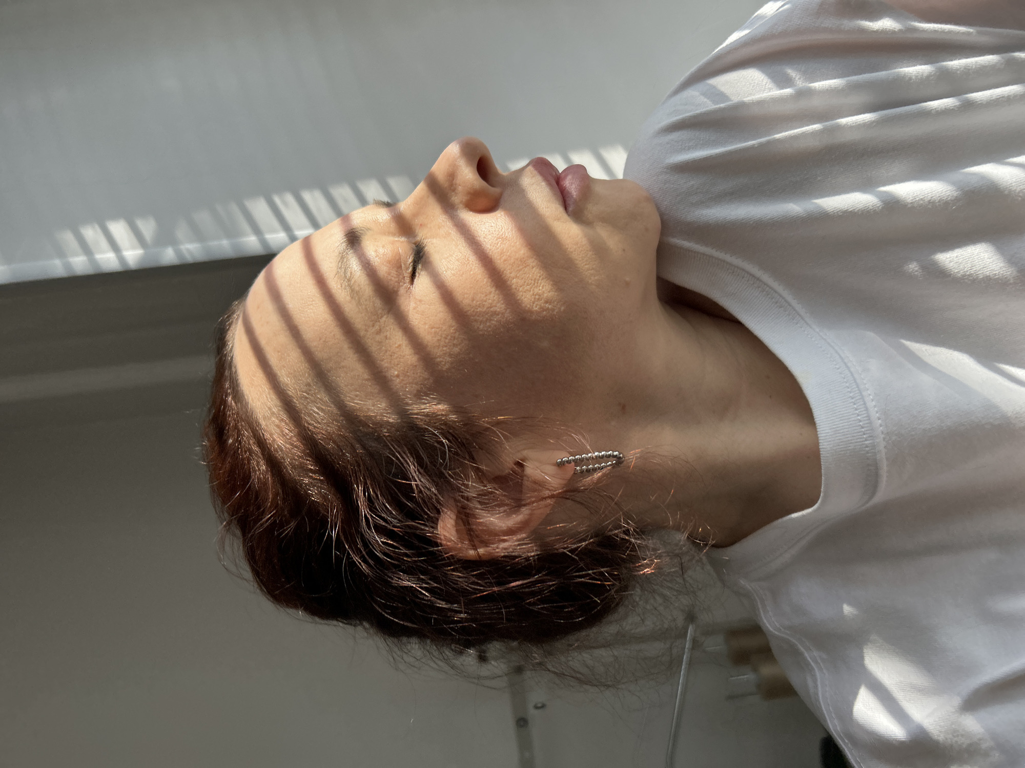 A woman with auburn hair and a white t-shirt is lying on her back, with her head tilted back. Light from a window with blinds is casting striped shadows across her face. She is wearing a silver ear cuff in her left ear. The location cannot be confirmed as Chaikovsky, Russia with the provided information.