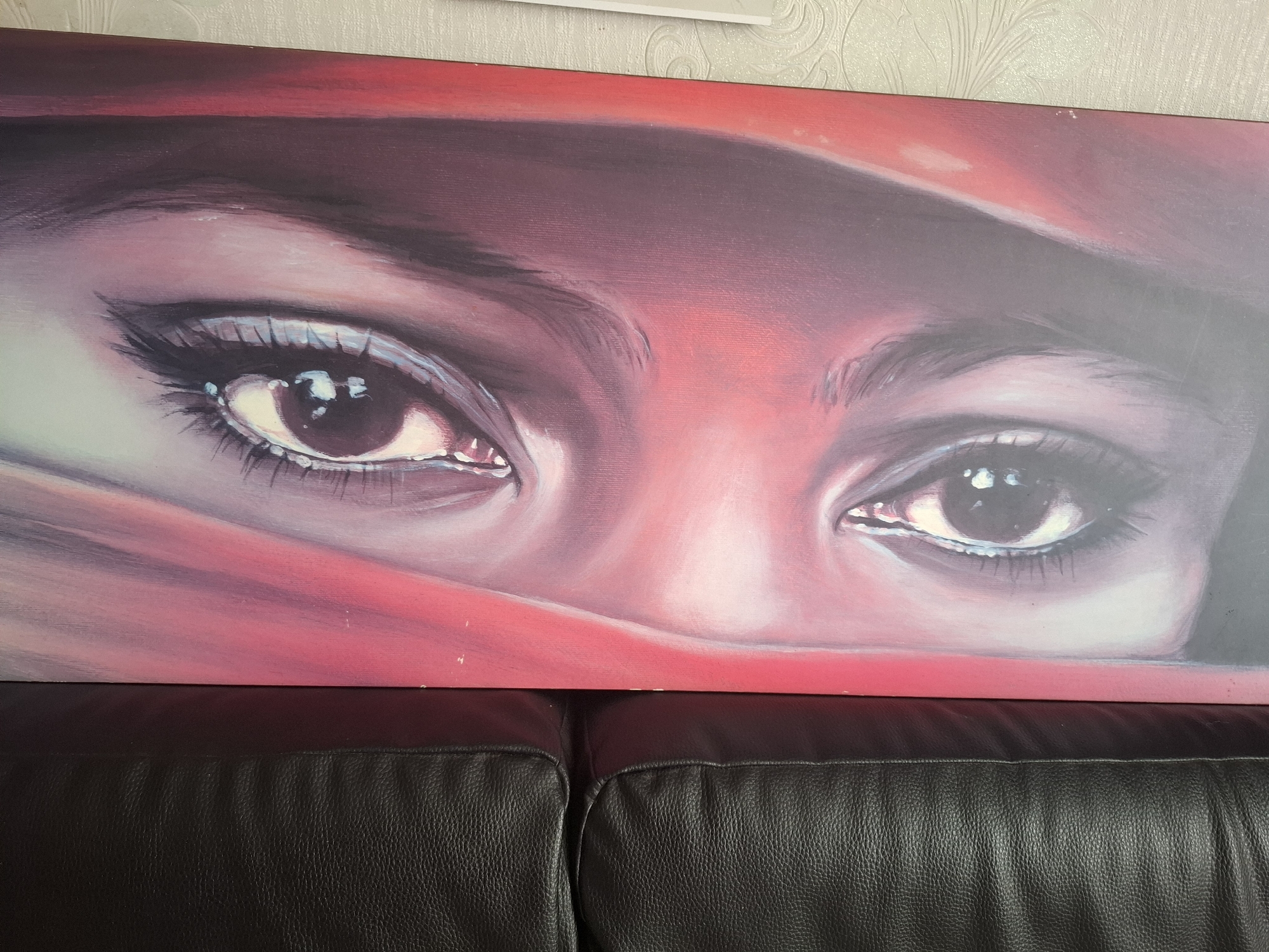 The image displays a close-up painting of a person's eyes, partially covered by a draped red fabric. The style is realistic, with detailed rendering of the eyelashes, eyelids, and the reflections within the irises. The background of the painting transitions from dark purple to bright red. Below the painting, a dark, textured surface, likely a leather sofa, is visible. The painting appears to be mounted on a wall. The image itself was taken in Soisy-sous-Montmorency, France, in a domestic setting.