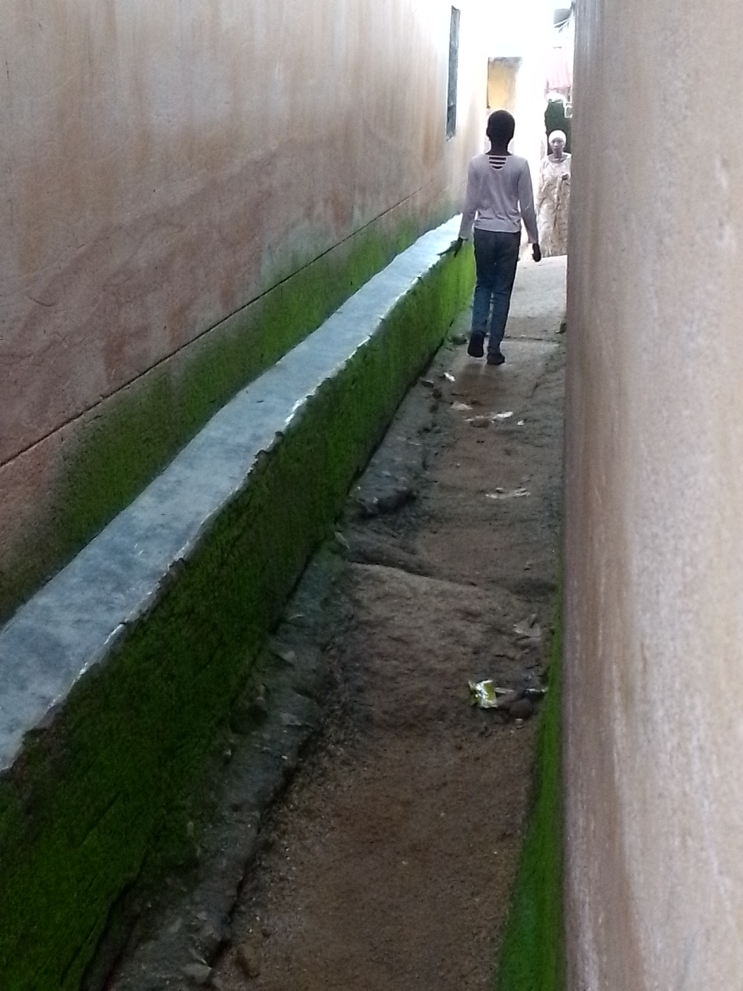 A person is walking away from the camera down a narrow alleyway. The alley is bordered by two tall, textured, beige walls. To the left, a concrete channel runs along the wall, filled with dark, damp soil and vibrant green moss. The person is wearing a light-colored, long-sleeved shirt with a striped neckline and dark pants. They are walking on a dirt path that slopes slightly downward. In the background, another person, wearing a patterned dress and a headscarf, is visible in the distance. The alley opens up to a bright, outdoor space at the far end.