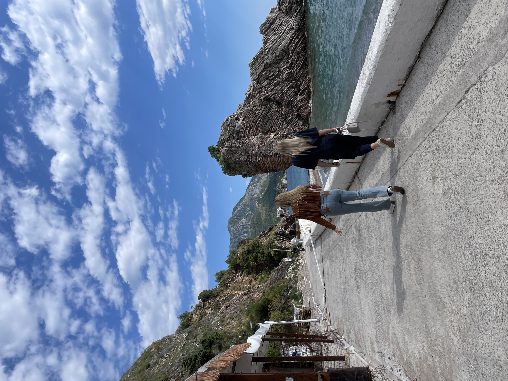 Two women stand on a concrete walkway bordered by a low, white wall overlooking the sea. The woman closest to the viewer wears light blue jeans with star embellishments on the rear pockets and a brown fringed jacket. The other woman is dressed in all black. The sea is on the right, with a rocky outcrop jutting out. On the left, the land rises sharply, covered in greenery and a building with a partly visible white roof. The sky is blue with scattered clouds. The location is Rafailovići, Montenegro.