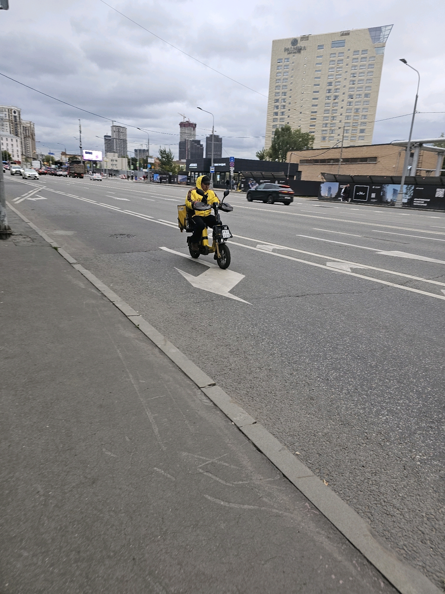 A person in a yellow hooded jacket and black pants is riding a yellow electric scooter down a multi-lane road in Moscow, Russia. The scooter has a yellow cargo box attached to the rear. The rider is wearing a black cap. In the background, there are several tall buildings, including a distinctive modern skyscraper with a slanted roof that appears to be the Neva Towers complex. There are also cars on the road, traffic lights, street lamps, and utility wires. The sky is overcast with gray clouds. The road is paved and marked with white lane lines and arrows. The scene suggests an urban environment with ongoing construction, as indicated by a building with scaffolding and cranes in the distance.
