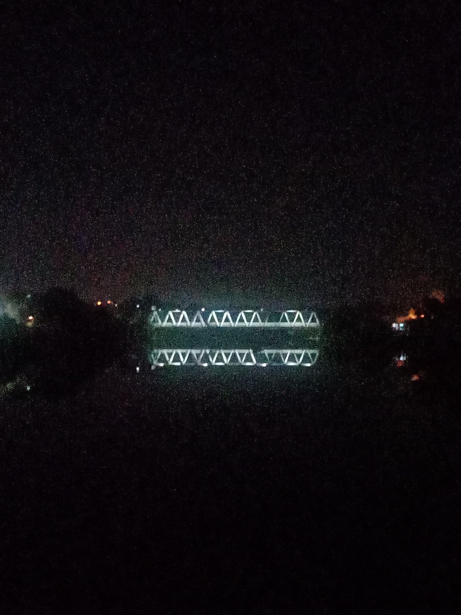 The image captures a night-time scene of an illuminated bridge spanning across a calm body of water in Lhokseumawe, Indonesia. The bridge structure, likely a truss bridge, is prominently lit with white lights that accentuate its geometric design, featuring a repeating pattern of triangles. These lights form two parallel horizontal bands across the center of the frame, with the upper band representing the actual bridge and the lower band being its clear reflection on the water's surface.
The surrounding environment is shrouded in darkness, emphasizing the brightness of the bridge. On either side of the water, distant, scattered lights can be seen, hinting at the presence of a nearby town, road, or settlement. Faint outlines of land or foliage are barely discernible against the dark background on the banks.
No specific human activity or individuals are visible in the photograph. The notable details include the time of day being clearly night, the serene appearance of the water creating a near-perfect reflection, and the architectural focus on the bridge. The image exhibits a grainy texture, typical of photography in low-light conditions. There is no visible text present in the image.