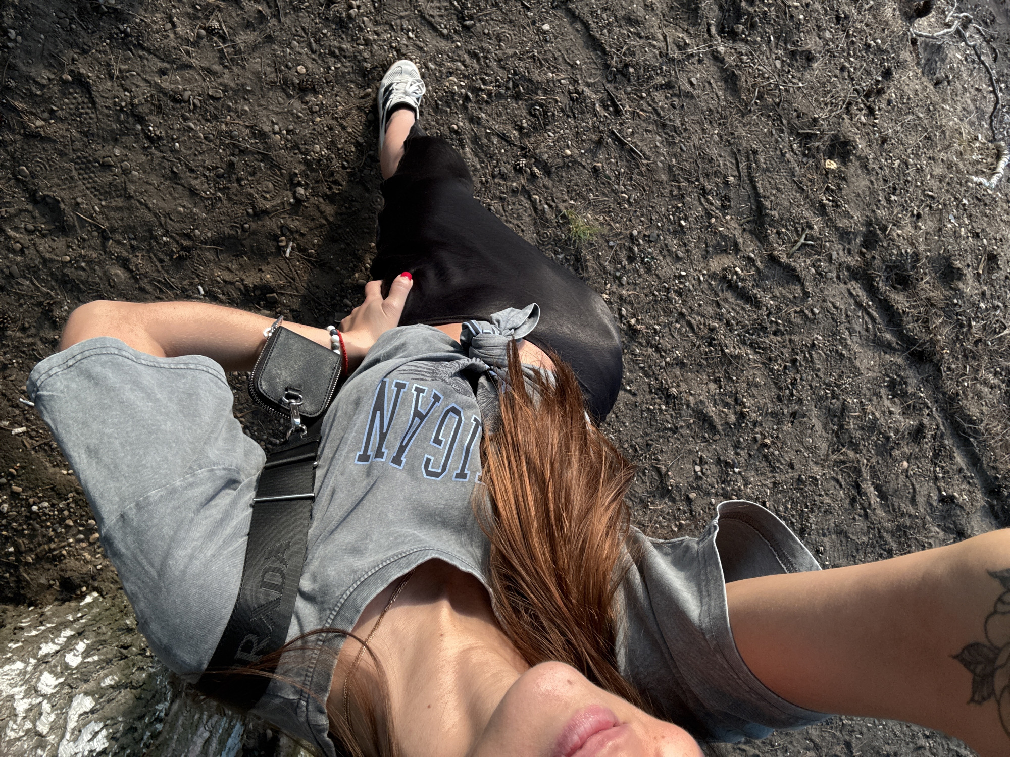 The media file shows a person lying on the ground, viewed from above. The subject has long, reddish-brown hair and is wearing a grey t-shirt with "NYGN" printed on it, grey pants, and white sneakers with black stripes. A black purse with a silver clasp is slung across their chest. The ground is dark and appears to be dirt with small stones and some dried vegetation.
The lighting suggests it is daytime, with sunlight casting shadows. The scene appears to be outdoors, possibly in a natural or unpaved area. The person is looking upwards and slightly towards the camera, with their mouth slightly open, giving a relaxed or candid impression. The person's right arm is bent at the elbow, resting on their head, and their left arm is partially visible near the purse. A tattoo is visible on their right arm. The overall mood is casual and relaxed, possibly a moment of rest or leisure.