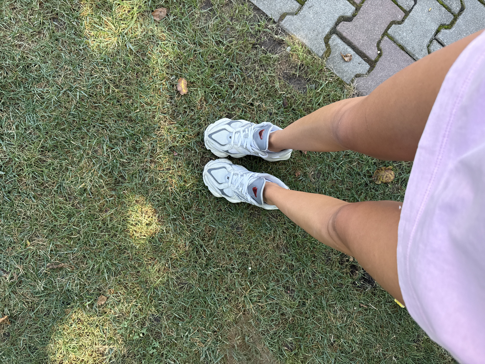 A first-person perspective shot shows a person standing on a lawn with a paved pathway to the right. The person is wearing white athletic shoes and a light pink t-shirt. The lawn is green with some dry patches and fallen leaves. The pathway is made of interlocking grey pavers. The scene appears to be outdoors, possibly in a park or backyard, in Cârțișoara, Romania.