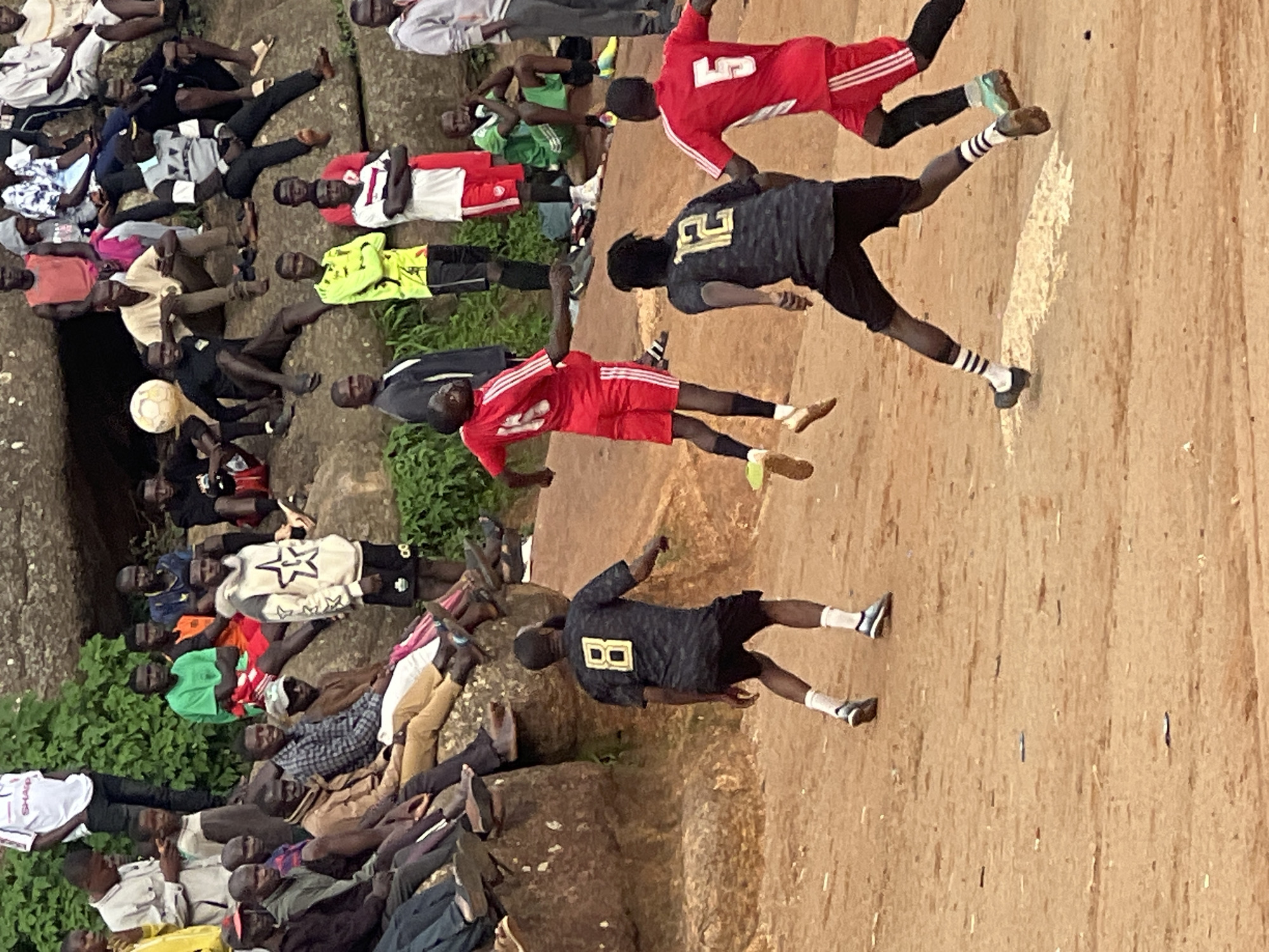 This media file captures a group of people, predominantly young men, engaged in a soccer match. The scene is set outdoors, on a rough, reddish-brown dirt field, possibly in Jos, Nigeria, given the context. Several individuals are actively playing soccer, kicking and running, while others are gathered on the sidelines, watching the game. Some are seated on a rocky outcrop covered with sparse vegetation, while others are standing or sitting on the ground.
The players are wearing various athletic attire. One player in the foreground is in a red jersey with white stripes on the sleeves and red shorts. Another is in a dark jersey with the number "12" in gold on the back and black shorts. A player in a lime green jersey and black shorts is also visible. Another player wears a red jersey with white stripes and red shorts. The spectators are dressed in casual clothing, including shirts, t-shirts, and some traditional Nigerian attire.
The lighting suggests it is daytime, likely mid-morning or afternoon, with no strong indications of specific weather conditions beyond it being outdoors. The mood appears to be one of casual recreation and community engagement. The presence of a soccer ball in the image clearly indicates the sport being played. The background shows a rocky, somewhat uneven terrain with patches of green vegetation, typical of an outdoor setting in a potentially arid or semi-arid region. The image is oriented vertically, with the action appearing to move from the bottom to the top of the frame.