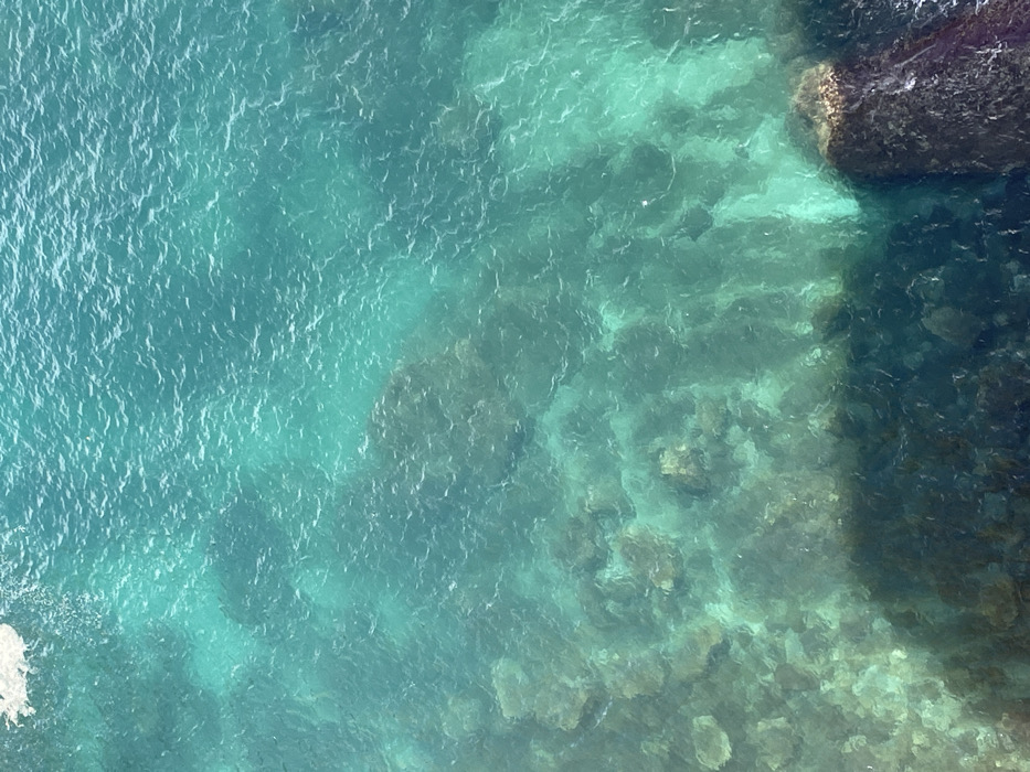 The image depicts an overhead view of clear coastal water in Budva, Montenegro. The water exhibits a range of turquoise and teal hues, with small, irregular ripples visible on its surface and a patch of white foam at the upper-left. Submerged below the transparent water, the rocky seabed is discernible, composed of irregularly shaped rocks appearing dark brown, green, and grey. A prominent dark rock formation, likely a section of a cliff base or large boulder, occupies the lower-right portion of the frame, extending into the water. A large, dark shadow is cast across the lower-left section of the water and submerged rocks. No human figures, marine vessels, or specific marine organisms are identifiable. The scene depicts a natural, rocky shallow water marine environment.