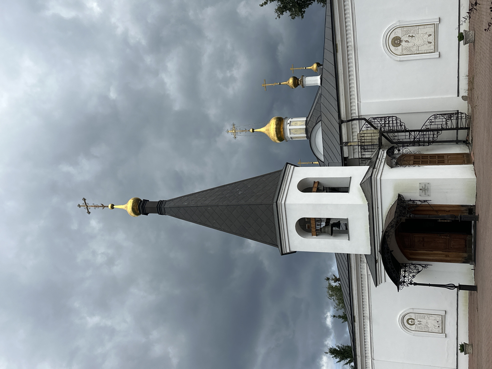 The image shows the exterior of a white church in Podolsk, Russia. The church has a tall, dark, conical roof that extends into a golden dome topped with a cross. There is a bell tower with three bells. Other golden domes with crosses are visible on the roof. The church walls are white, with two mosaic icons on the exterior. There is a wooden door with an ornate metal awning above it. A spiral staircase made of black metal is attached to the side of the building. The sky is overcast with dark clouds. The ground in front of the church is paved with red bricks. Trees are visible in the background. There are no people in the image.