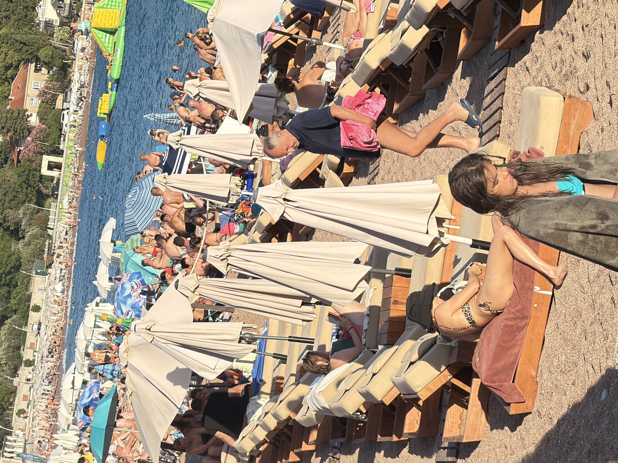 The image depicts a crowded beach scene in Petrovac centar, Montenegro, under a bright sky. Numerous beachgoers are scattered across the sand, with many relaxing on lounge chairs positioned under beige umbrellas. Some individuals are seated on the chairs, while others are partially submerged in the clear blue water. A few are engaged in swimming or playing in the sea, where inflatable water attractions are visible. A man in a dark blue polo shirt and dark shorts is walking across the beach, carrying a pink towel. In the foreground, a woman wearing a leopard print bikini and a black sun hat is lying on a lounge chair. Another woman, wrapped in a dark towel with her wet hair visible, sits in the foreground. The background shows a tree-lined promenade and buildings. The overall atmosphere is that of a busy summer day at the beach.
