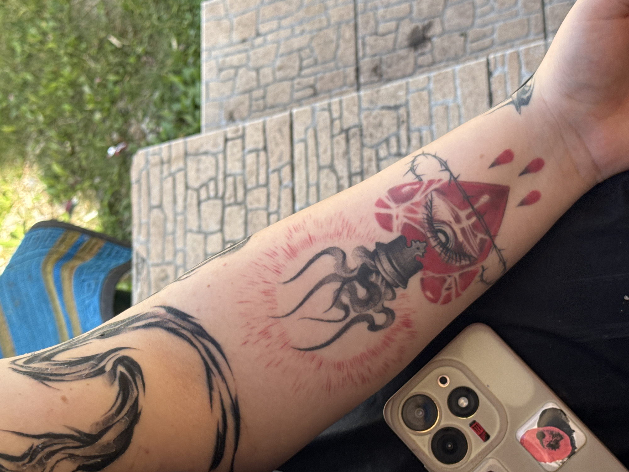 The image shows a close-up of a person's arm, which is adorned with several tattoos. The main subject is the arm with tattoos, displaying a chess piece, possibly a king, with smoke-like tendrils emanating from it, surrounded by a red aura. Above this, there is a heart-shaped design with an eye and barbed wire. Further up the arm, black abstract shapes resembling waves are visible. In the foreground, a corner of a smartphone with a light-colored case and multiple camera lenses is visible. A sticker with a red floral design is partially visible on the phone case. The arm appears to be resting on dark fabric. The background is out of focus, showing some greenery and textured stone steps, suggesting an outdoor setting. There are no discernible words or text in the image. The lighting suggests it is daytime.