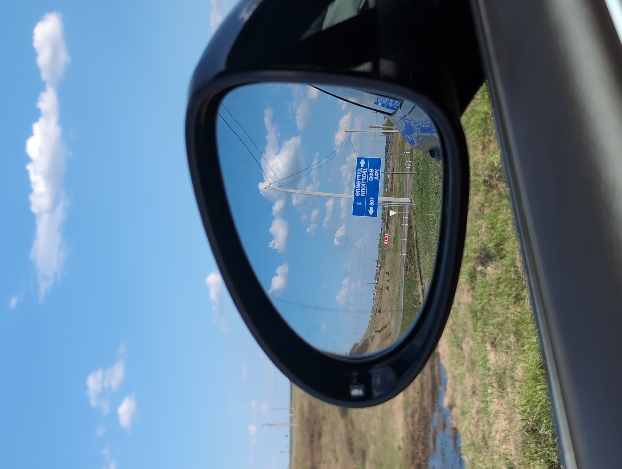The image shows a car's side mirror reflecting a view of a rural area near Такарликово, Russia. The mirror reflects a blue sky with white clouds, a road sign indicating directions to different locations, power lines, a grassy field, and a distant horizon. The sign appears to display Cyrillic text, providing place names and directional information. The reflection is somewhat distorted by the curve of the mirror. The mirror itself is black and attached to the car. The focus is on the reflection within the mirror, suggesting it is the primary subject of the image.
