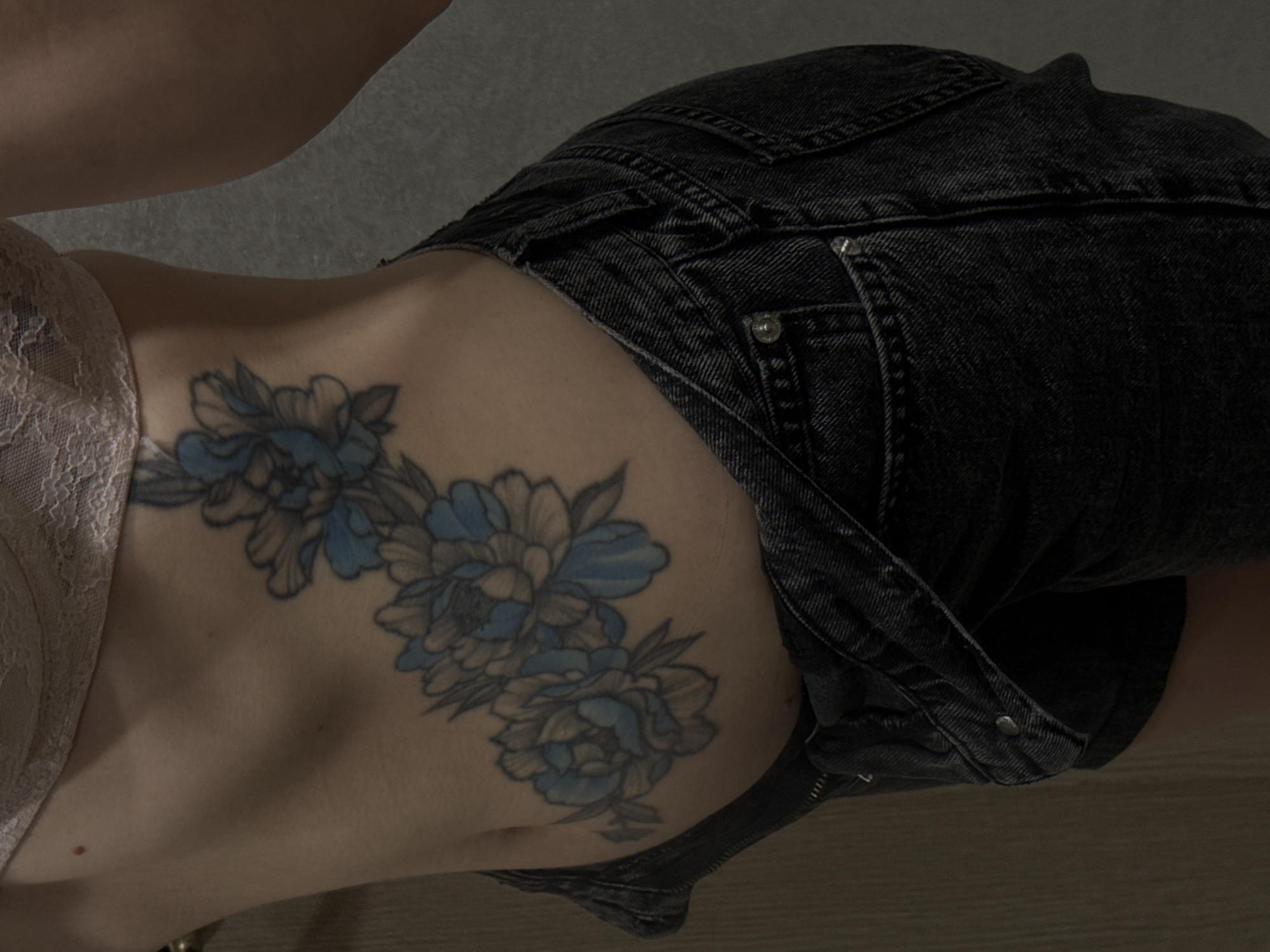 The image is a medium shot of a person's torso, from the waist up, and it is not possible to determine the location of the city of Moscow, Russia, from the image. The person is wearing a light-colored lace bra and dark denim overalls that are unzipped, revealing part of their midriff. A tattoo of blue and grey flowers is visible on the person's left side, extending across their waist and lower ribs. The background is a plain, textured grey wall on the right and a textured light-brown surface on the left, possibly a door or wall. The lighting is dim, with shadows obscuring some details.
