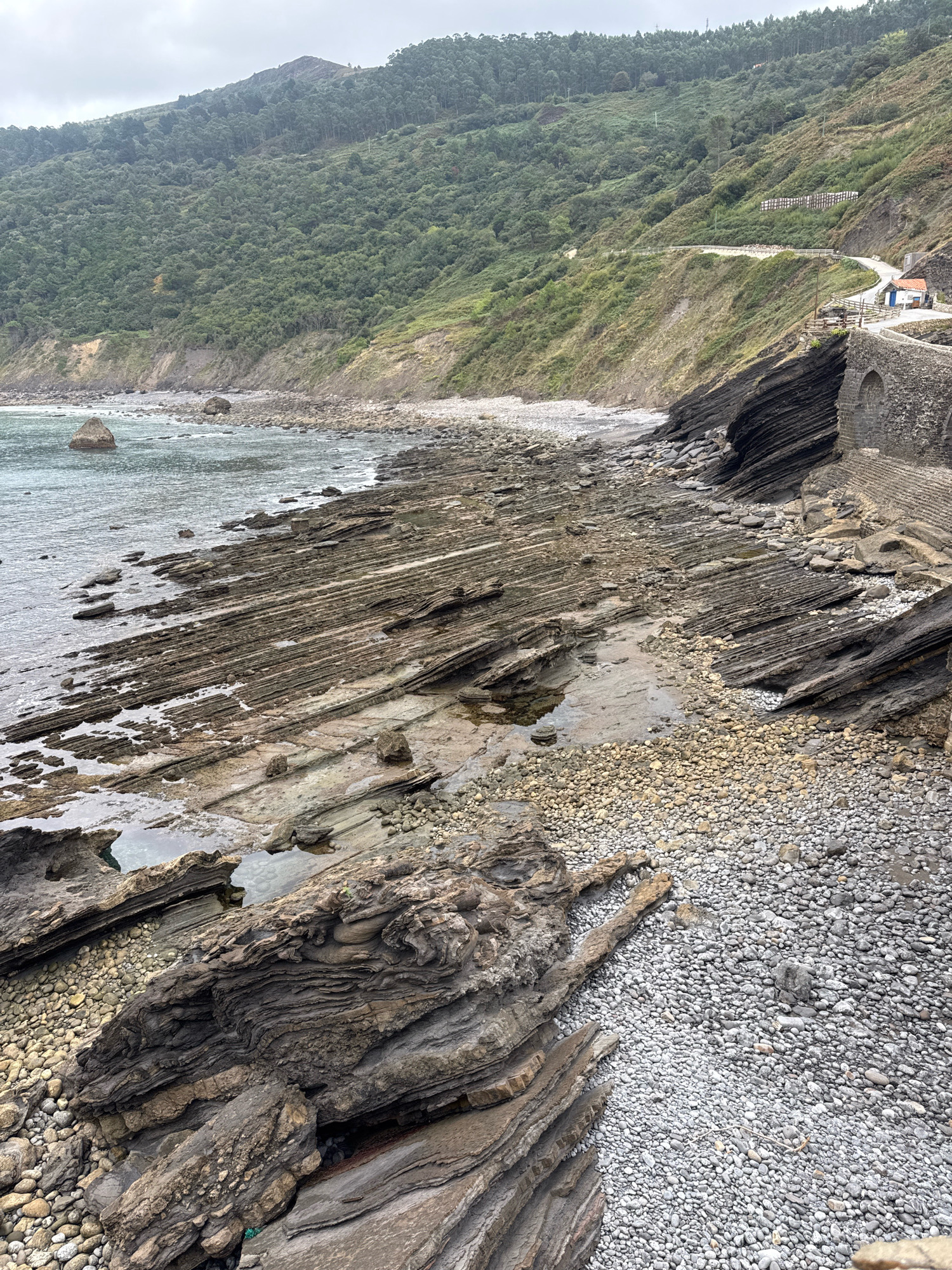 This is a high-angle medium shot of the rocky shoreline near the San Juan de Gaztelugatxe sanctuary in Bakio, Spain. The scene is dominated by layered, dark gray and brown rock formations that jut out into the choppy, gray-blue water. The rocks show evidence of erosion, with distinct horizontal striations and uneven surfaces. The shoreline is also covered in a mix of smaller, rounded pebbles and scattered larger rocks. In the background, a steep, green hillside covered in dense trees rises sharply. A winding paved road with a white railing is visible ascending the hillside, leading to a small white building with a red-tiled roof. A stone bridge structure with arched openings is visible on the right side of the frame, partially obscuring the view of the road and building. The sky is overcast and a pale gray. There are no people or animals visible in the image.
