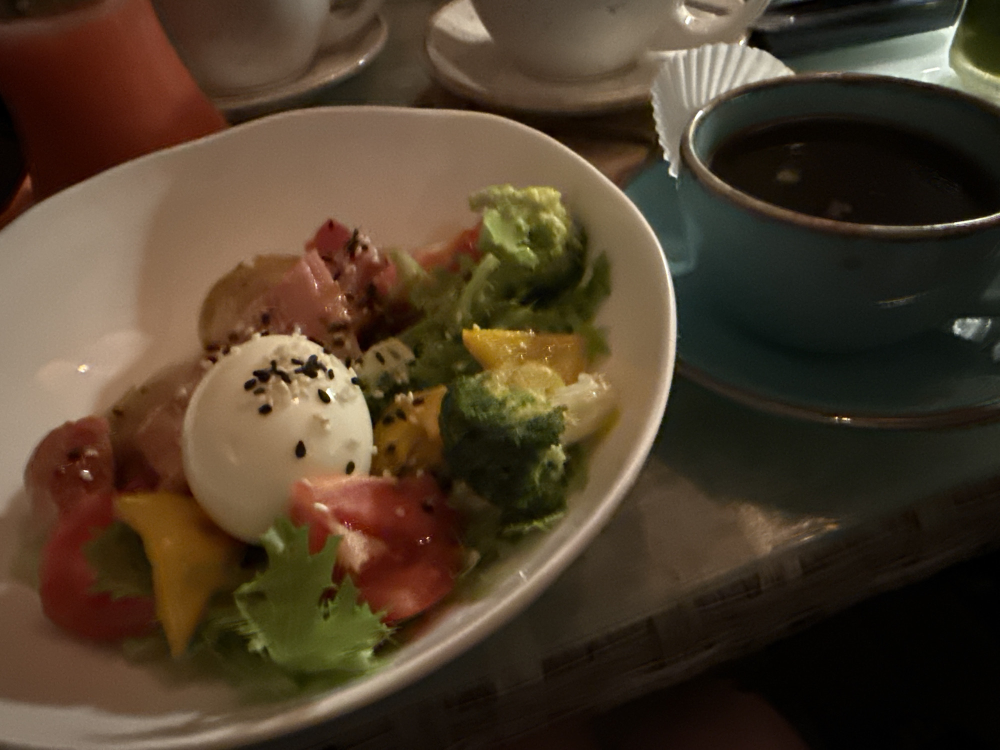 A close-up shot captures a white bowl filled with a colorful salad, featuring a hard-boiled egg topped with black sesame seeds, broccoli florets, tomatoes, and yellow bell pepper. Beside the bowl, a blue ceramic cup holds dark liquid, likely coffee. The scene is set on a table with a textured surface, hinting at a casual dining environment, potentially in Feodosia, Ukraine.
