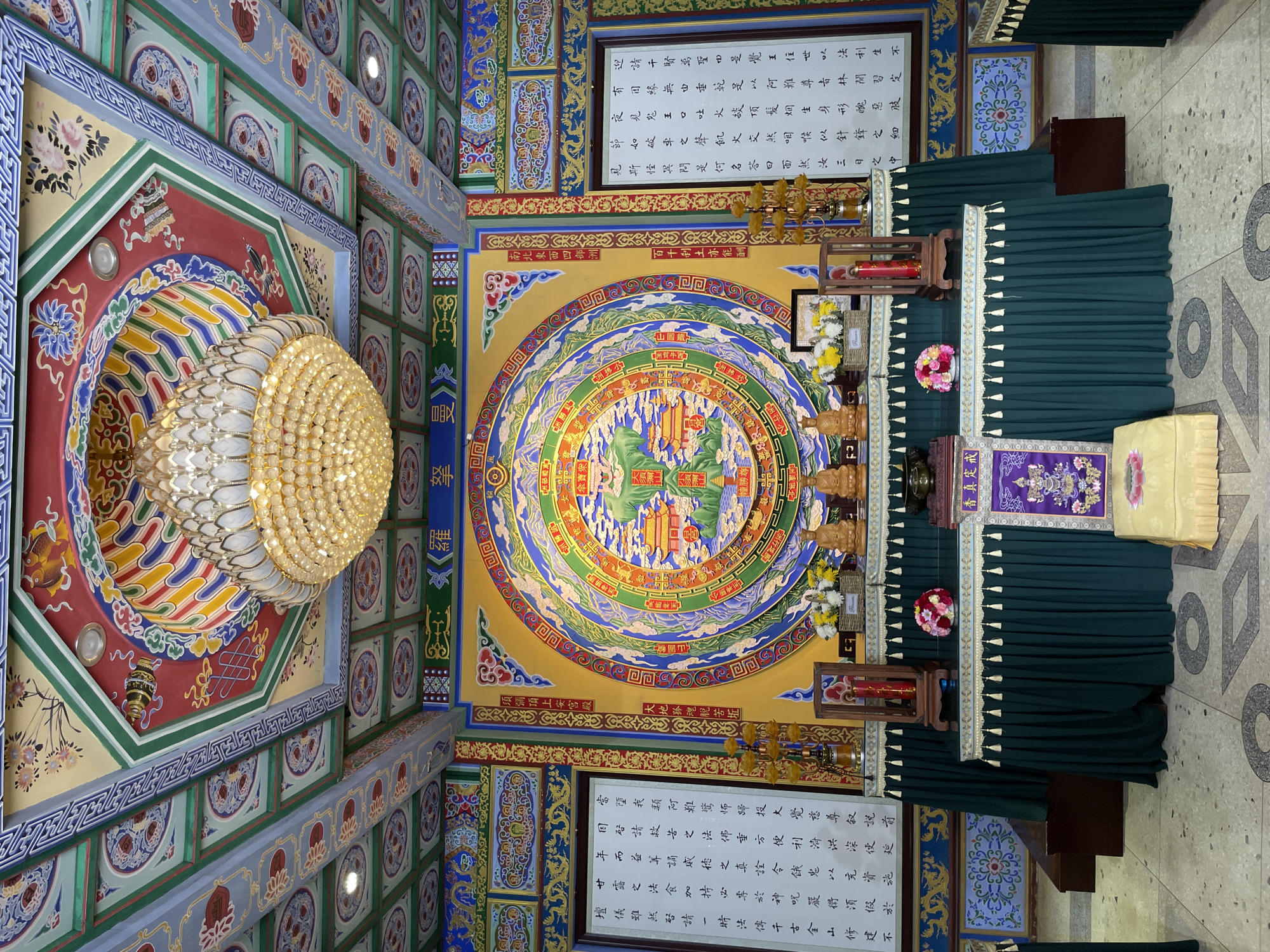 The image is an interior shot of a room decorated with intricate traditional Chinese designs, likely a temple or religious site located in the Yanta District of China. The walls and ceiling feature a variety of patterns, including circular motifs, floral designs, and calligraphy. A large, ornate chandelier hangs from the ceiling, providing illumination. The room also contains a draped altar with what appear to be religious artifacts.