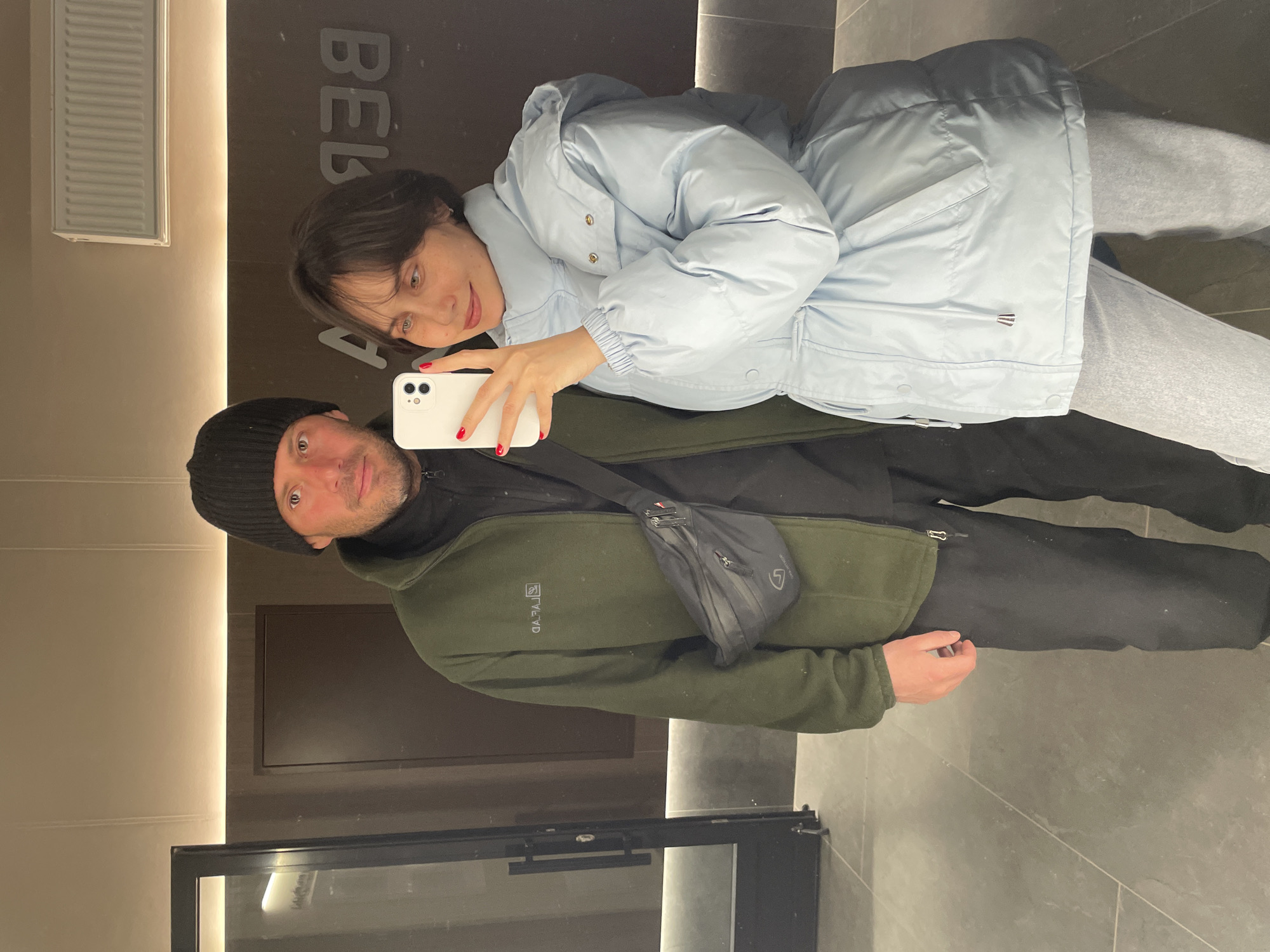 A man and a woman are taking a selfie inside a building. The woman is holding a white iPhone and smiling at the camera. She is wearing a light blue puffer jacket and light-colored jeans. The man is wearing a black beanie, a dark green fleece jacket, and black pants. He has a black crossbody bag. They are standing in front of a dark-colored wall with text in Cyrillic script. A door with a black frame and glass panels is visible in the lower-left corner. The floor is tiled with large gray tiles. The location is Izhevsk, Russia.