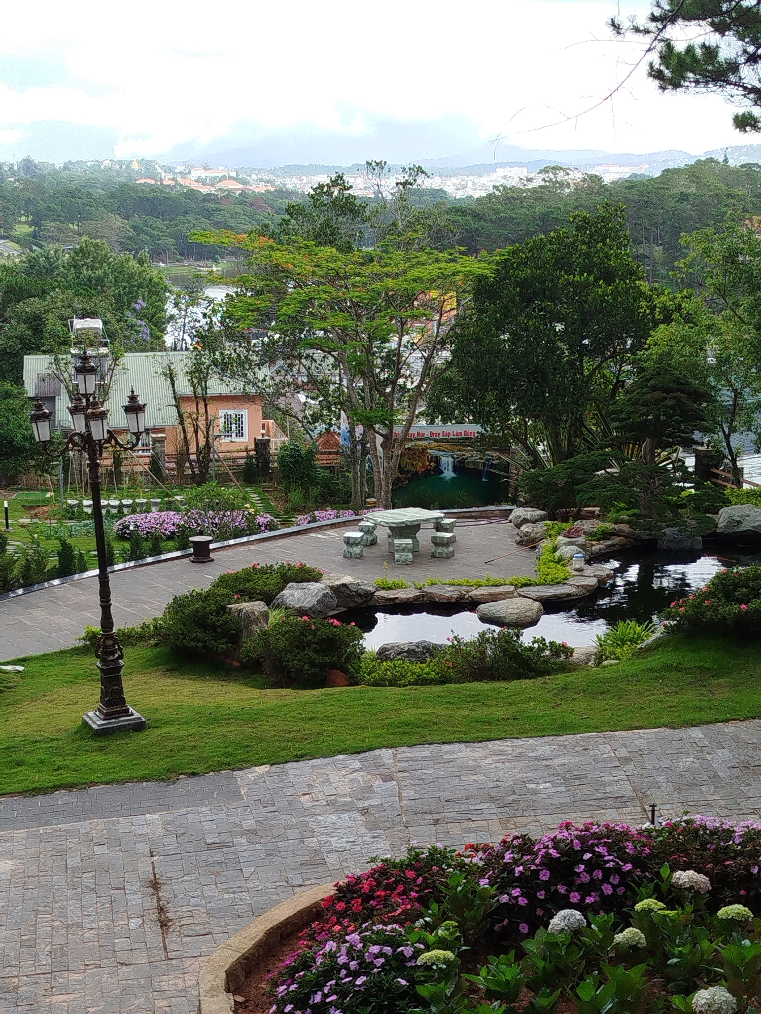The image shows a scene in Da Lat, Vietnam, likely taken from an elevated position. The foreground features a paved walkway with flowerbeds containing red, pink, purple, and white flowers. To the left, a tall, ornate lamppost stands on a grassy area. Beyond the walkway, there is a pond surrounded by rocks and greenery, leading to a patio area with a stone table and chairs. Behind the patio, there's a backdrop displaying a waterfall scene and Vietnamese text. Further in the background, there is a building with a peach-colored exterior and green roof, along with other trees and greenery. In the distance, a cityscape is visible under a cloudy sky. The overall impression is of a peaceful and well-maintained garden or park area.