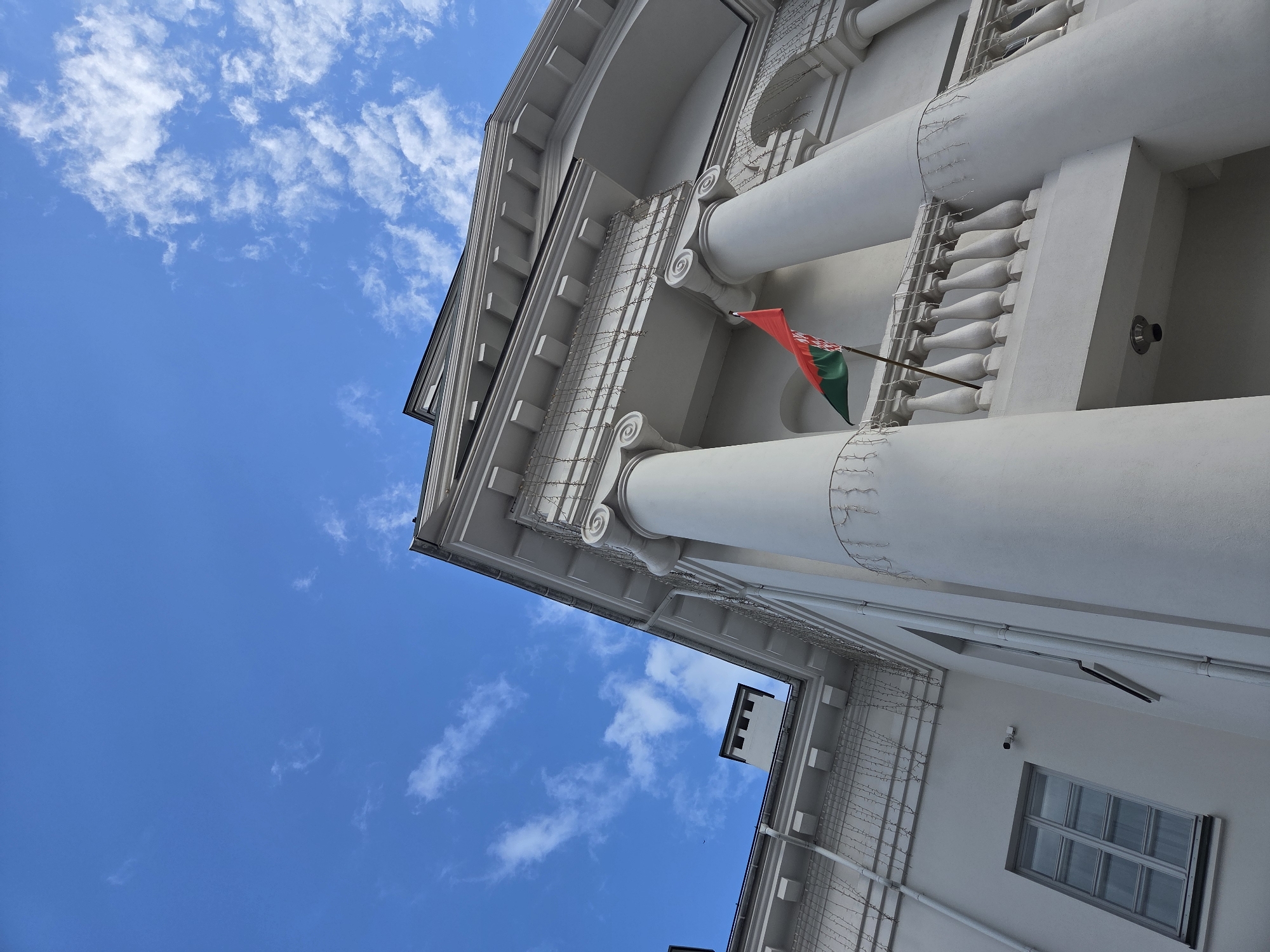 A low-angle outdoor shot shows the white facade of a building with a classical architectural style, featuring large columns and a decorative railing on a balcony. A flag, red on top and green on the bottom with a white pattern on the green section, is flying from the balcony. The sky above is a clear blue with scattered white clouds. There is a window with multiple panes on the lower left of the building, and a security camera is visible above it. A chimney with square openings is seen on the roof. String lights are draped along the upper edge of the building and around the columns.