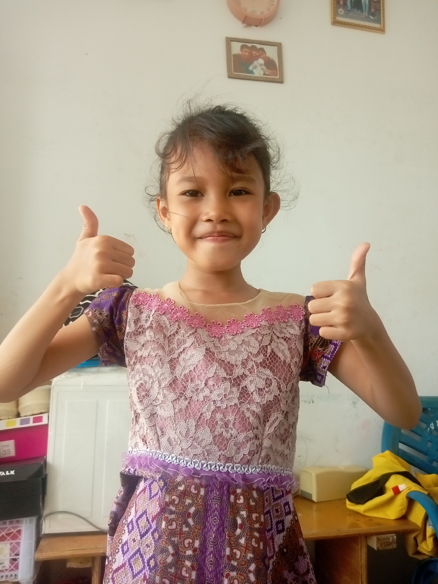 A young girl with dark hair pulled back and wearing a purple and pink lace dress stands in front of a white wall. She smiles and gives two thumbs up. Behind her, a clock and two framed pictures hang on the wall. To her left, a white refrigerator and some colorful boxes are visible. To her right, a wooden table holds a yellow bag and a blue chair. The scene suggests a casual indoor setting, possibly a home or a small shop, in Batam, Indonesia.