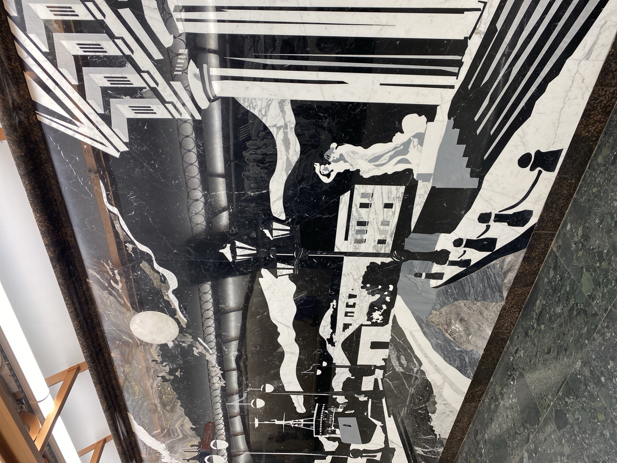This media file showcases a large-scale, intricate artwork made from inlaid marble or stone, predominantly in black, white, and various shades of grey. The artwork depicts a highly stylized and dynamic cityscape, likely a floor or wall mosaic in a grand public space in Saint Petersburg, Russia.
The style is evocative of Art Deco or Constructivism, characterized by bold geometric shapes, sharp angles, and strong contrasts. The scene features abstract representations of skyscrapers, buildings with distinct window patterns, and industrial structures. Interspersed throughout are flowing, wavy lines that could symbolize smoke, clouds, or water, and diagonal stripes that convey movement, light rays, or speed.
Subtle, highly stylized human figures are integrated into the design, including a profile of a face and abstract forms with elongated limbs, suggesting the bustling life within the city. A prominent white circular shape, possibly representing the moon or sun, is visible. Below it, intricate linear designs suggest elements like ships, bridges, or industrial machinery.
The monochromatic palette emphasizes the architectural forms and the interplay of light and shadow, with the natural veining of the marble adding texture and depth. The overall impression is a vibrant, modern metropolis, potentially celebrating industry and progress. The artwork is bordered by darker, granular stone in shades of brown and dark green, integrating it into the surrounding architectural elements. Reflections from overhead lights suggest an indoor setting. No visible text is present.