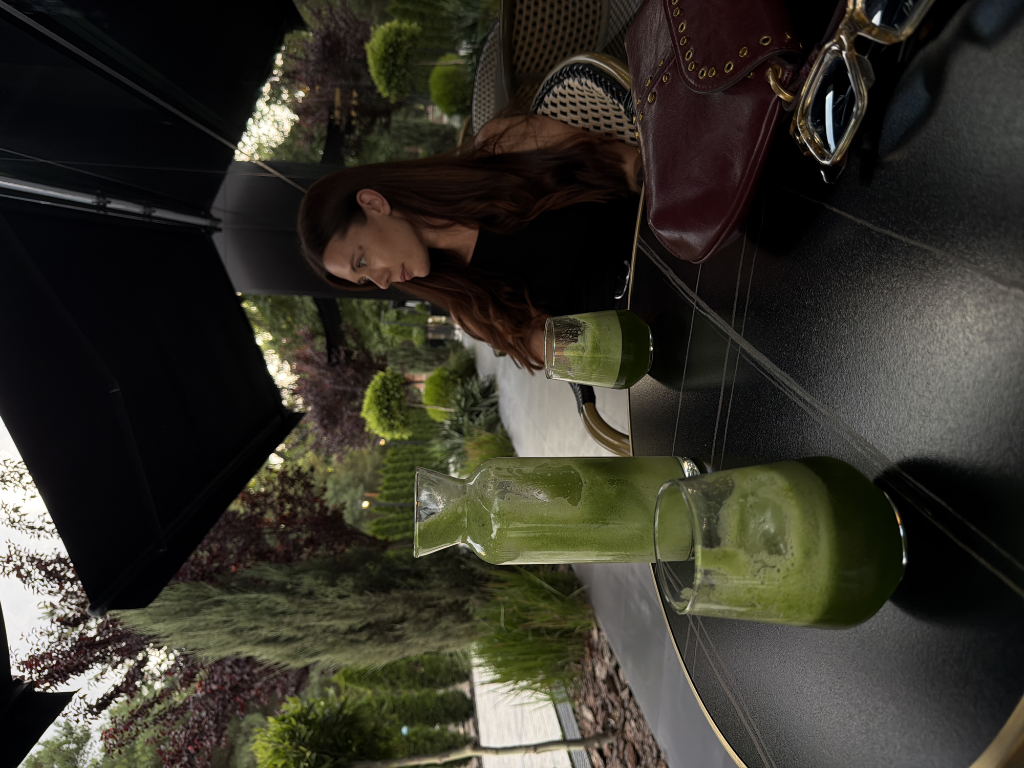 A medium shot captures a woman with long brown hair, wearing a black top, sitting at a black table on a patio. She is looking to her right, away from the camera. On the table in front of her, there is a glass carafe filled with a green beverage, two glasses of the same green beverage, a burgundy purse, and a pair of eyeglasses. The background shows a lush, green outdoor setting with trees and manicured bushes, suggesting a park or garden area. The patio appears to be covered by a dark awning. The scene implies a leisurely moment spent outdoors, possibly at a cafe or restaurant. There are no visible indications of the city of Bishkek, Kyrgyzstan.