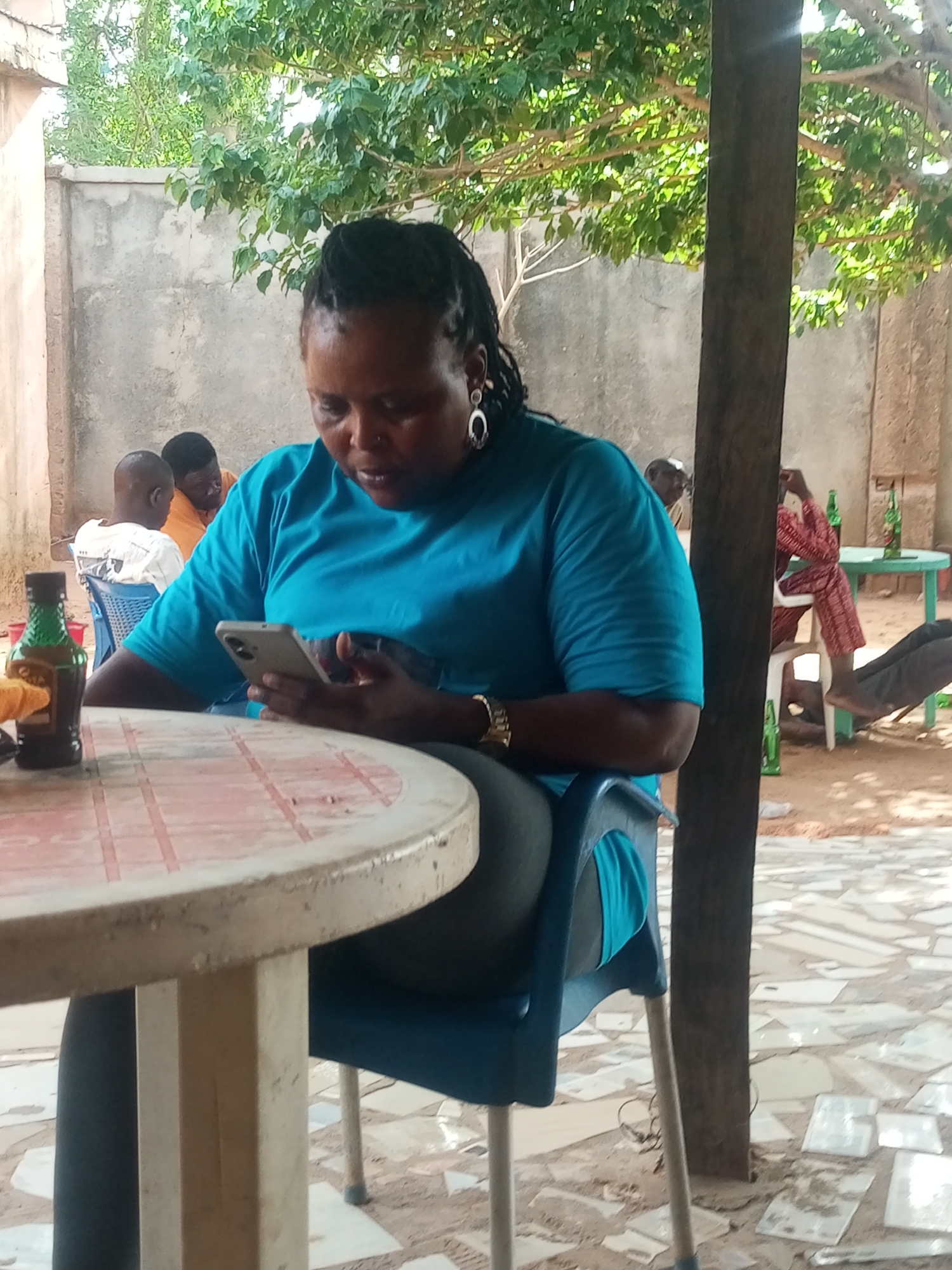 A woman with dark hair and a blue shirt is sitting at a round table outdoors, looking at her smartphone. She is wearing a watch and earrings. On the table, there is a bottle of liquid. In the background, other people are seated, and bottles are placed on a green table. The location appears to be a semi-outdoor setting with a wall and a tree providing shade. The ground is covered in light-colored stones. The city is unknown.