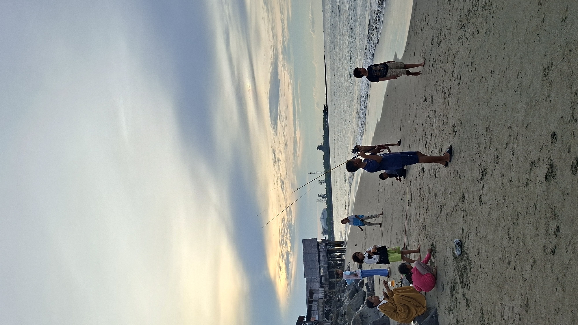 A group of people, predominantly children, are gathered on a sandy beach in Lhokseumawe, Indonesia, near a pier with buildings. The sky is overcast with patches of sunlight breaking through, creating a twilight atmosphere.
In the foreground, several children are engaged in various activities. Two children are sitting on the sand, one holding what appears to be a drink. Another child in a pink shirt is crouching and drawing on the sand. Two boys in the center are holding fishing rods, with one actively casting or reeling. To the right, another boy stands looking towards the sea. Further back, a woman in a hijab stands next to a group of children, one of whom is holding a white object. On the left side of the image, closer to the rocky shoreline and the pier, an elderly woman in a yellow dress is seated on a rock.
The environment consists of a sandy beach with gentle waves lapping at the shore. The pier structures are visible in the background, along with some vegetation and buildings in the distance. The overall scene depicts a casual evening gathering on the coast.