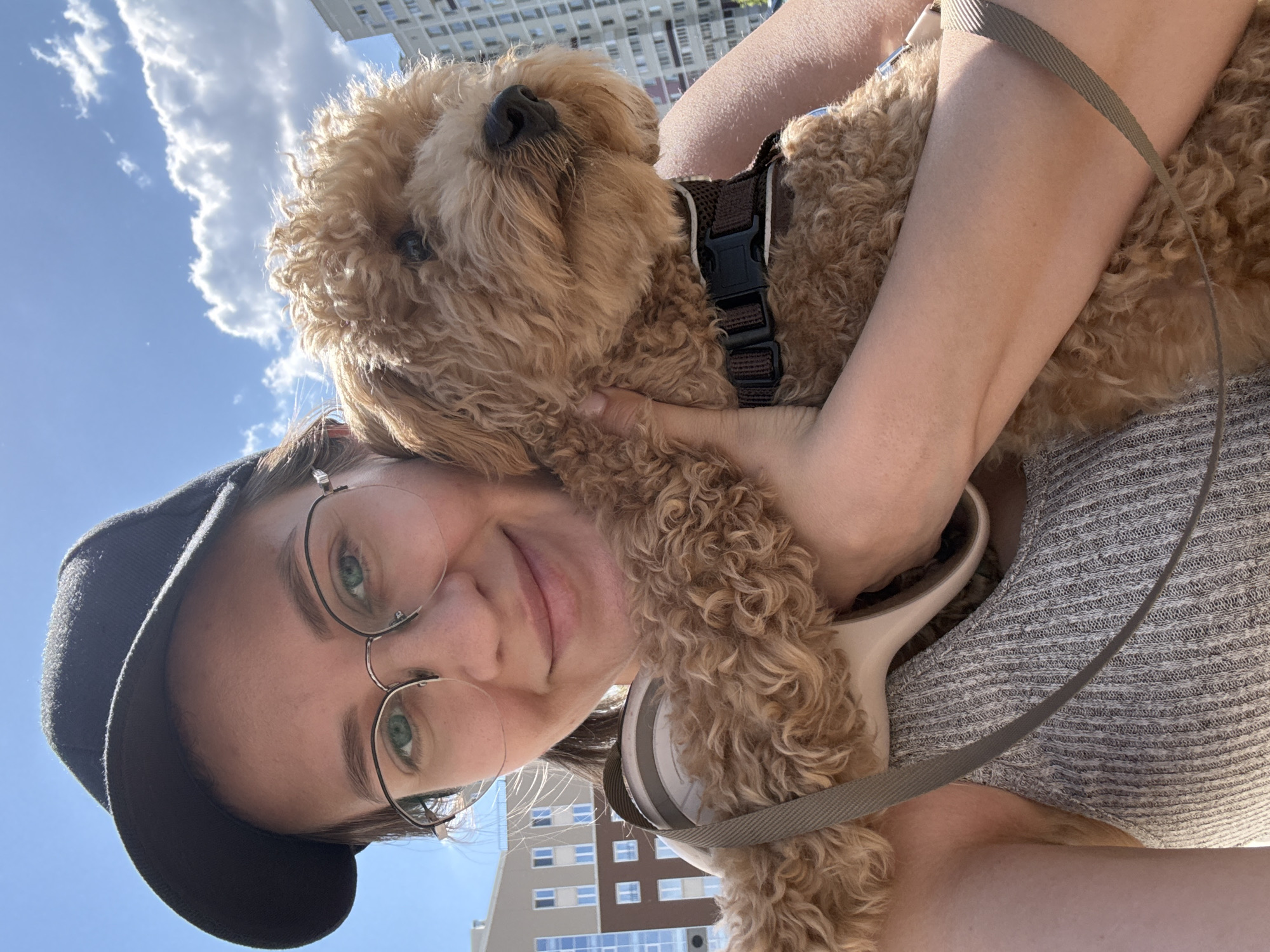 A woman in a black baseball cap and round wire-rimmed glasses holds a fluffy brown dog. She smiles at the camera. She wears headphones around her neck and a gray striped shirt. The dog wears a brown harness. A blue sky with white clouds is visible in the background along with the side of a multi-story building. The location is Izhevsk, Russia.