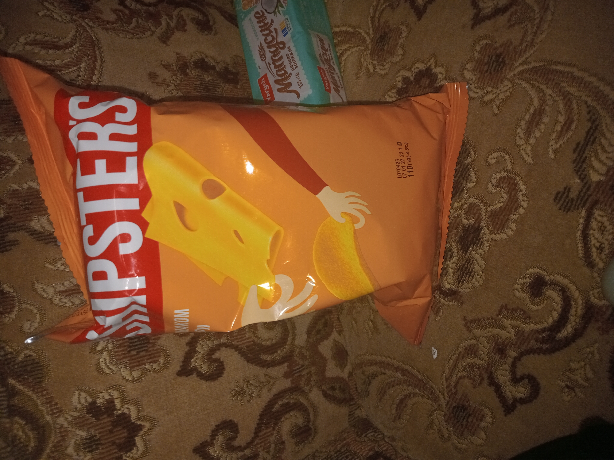 A bag of chips labeled "SNIPSTERS" with a cheese illustration is lying on a patterned brown fabric surface. To the right of the chip bag, a package of cookies labeled "Матусине" is visible. The lighting in the image is dim. The scene is indoors, on a couch or similar piece of furniture. No specific location in Perechyn, Ukraine, can be identified from the visual information provided.