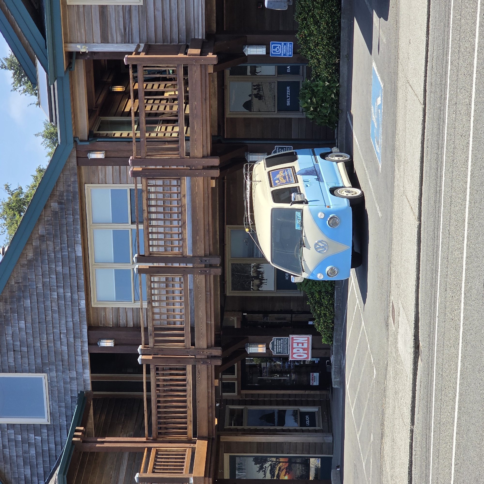 The image captures a front view of a blue and cream-colored Volkswagen Type 2 van parked in a lot in front of a wooden building in Cannon Beach, Oregon, United States. The building appears to be a commercial establishment, possibly a liquor store, with several windows and a covered walkway. A red "OPEN" sign is visible near the entrance. The van has a luggage rack on its roof and a sticker on its side depicting a coastal landscape. The parking lot has painted lines and a handicapped parking symbol. Behind the van, there are bushes and the building's facade. The sky is blue and clear.