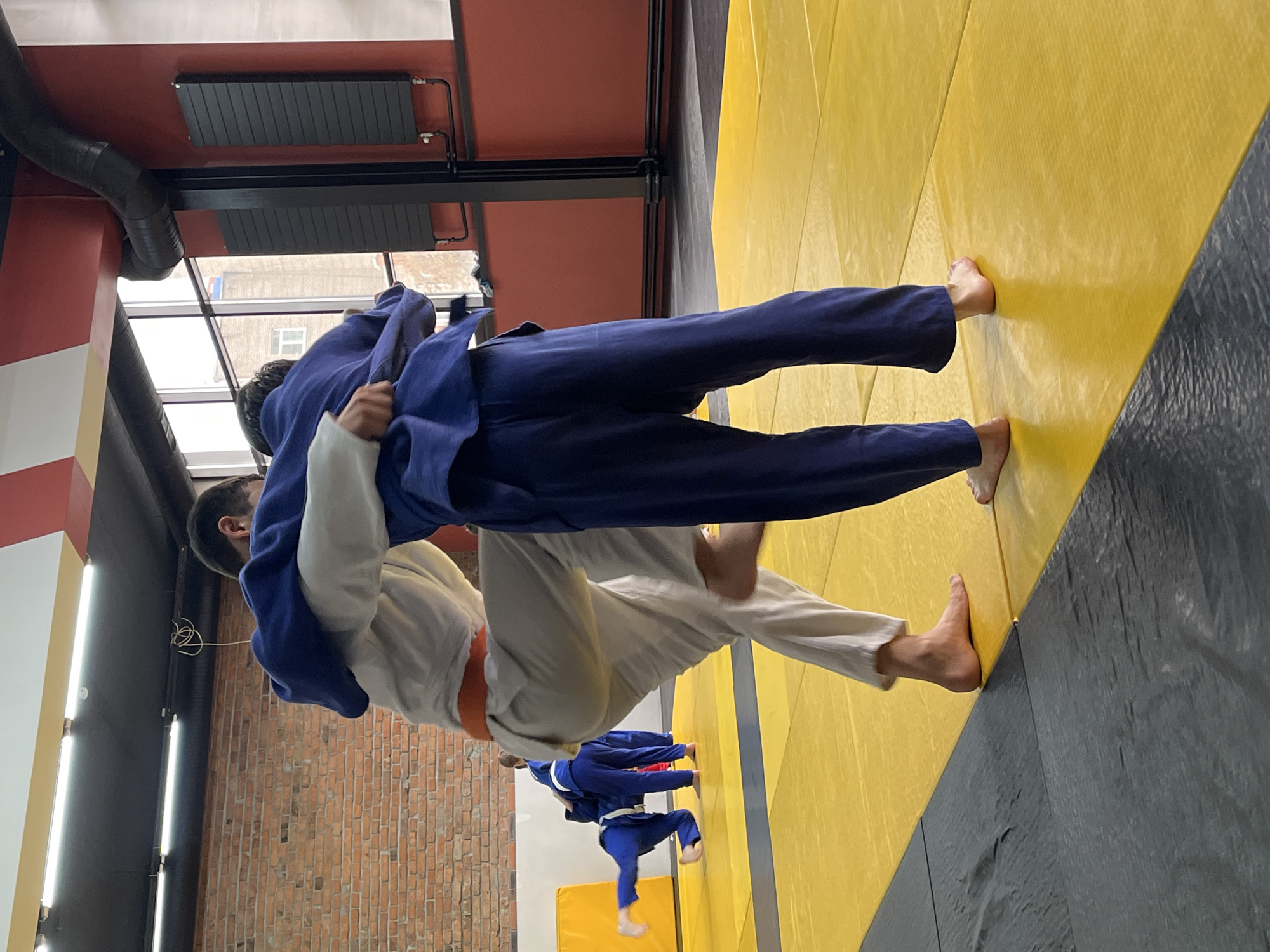 Two individuals are engaged in a judo match on a yellow and black mat inside a building in Krasnoyarsk, Russia. One individual wears a blue judogi and the other wears a white judogi with an orange belt. They are grappling with each other. Several other individuals in blue judogis are visible in the background. The building has brick walls and a window, with additional indoor fixtures.