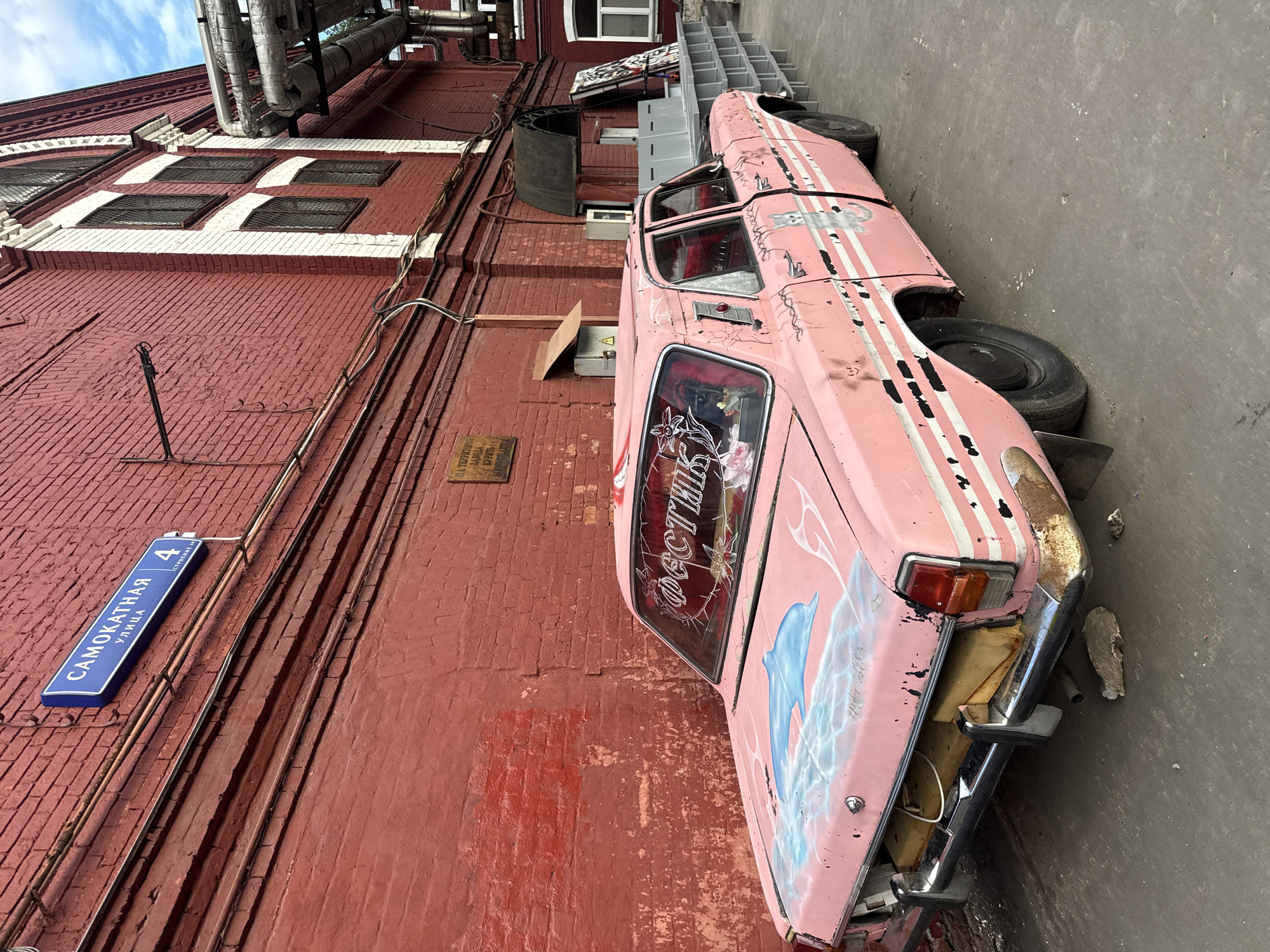 A pink, vintage sedan with rust and graffiti is parked in front of a red brick building. The building has pipes running along the exterior and windows with metal grates. A street sign on the wall reads "САМОКАТНАЯ УЛИЦА 4," indicating the location is Samokatskaya Street, building 4, in Moscow, Russia. The car's trunk is open, revealing yellow foam padding. The rear bumper is also rusted. The car has a dolphin painted on the hood and a cat painted on the side. The windshield has lettering that appears to say "ФЕСТИВАЛЬ" (Festival). The ground is a concrete surface.