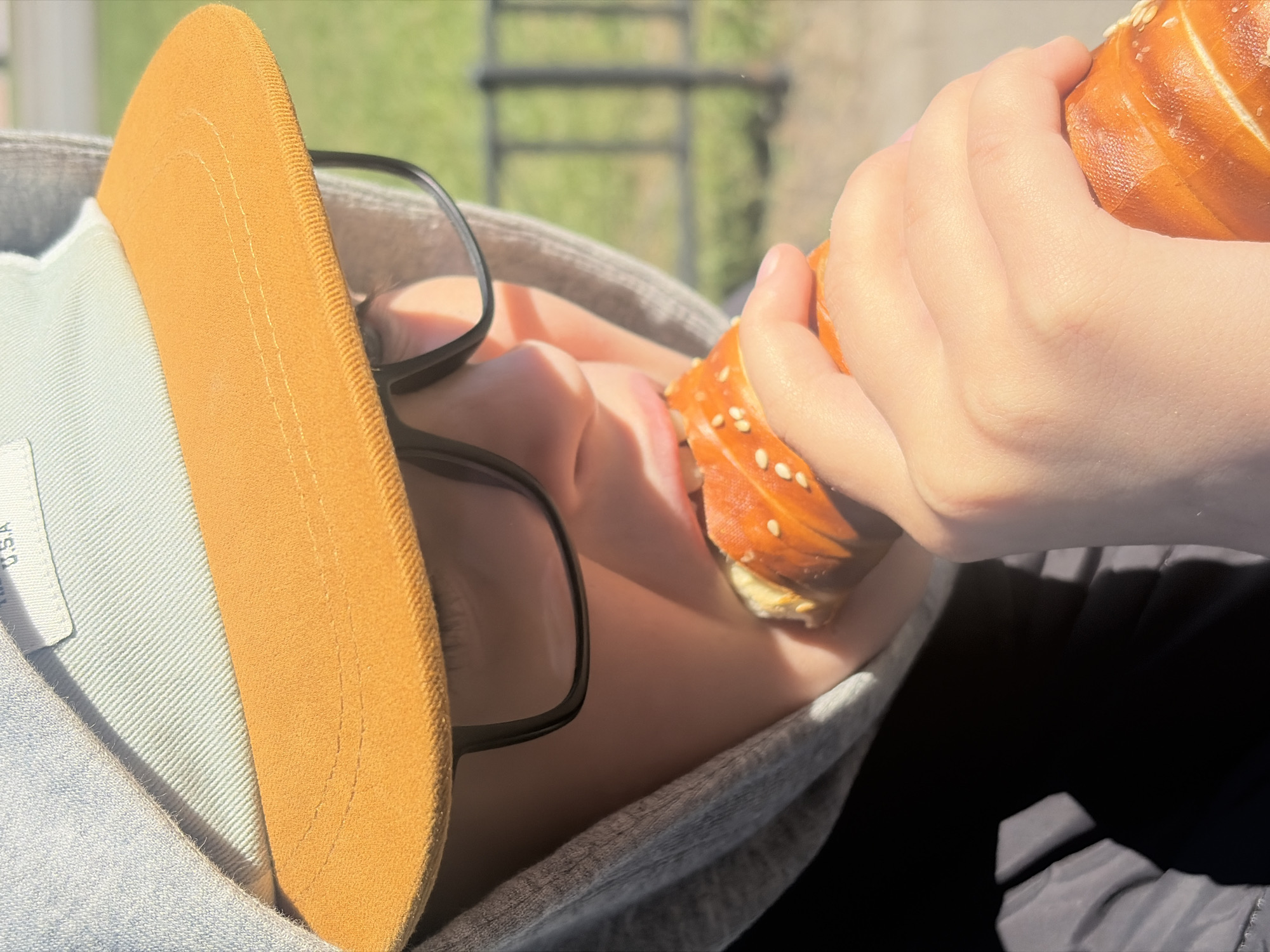 A close-up, high-angle shot captures a person, likely a child or young adult, eating outdoors in what appears to be a sunny setting in Saint Petersburg, Russia. The individual is seen from the nose up, wearing black-framed glasses, a grey hooded sweatshirt, and a light-colored cap with a prominent orange or tan brim. Their hands hold a golden-brown bread roll, possibly a pretzel, speckled with sesame seeds or salt, which they are actively biting into.
Bright sunlight illuminates the scene, casting strong highlights on the bread and the person's skin, and creating shadows, suggesting a clear, sunny daytime. The background is softly blurred, showing green foliage and what appears to be a metal fence or railing, indicating an outdoor park or recreational area. Faint text "U.S.A" is visible on the light-colored fabric of the cap near the brim. The overall impression is of a casual moment, enjoying a snack outdoors.