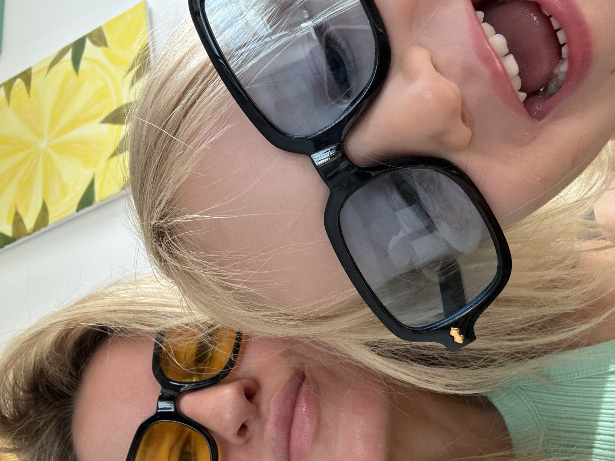 The image is a close-up selfie taken by two individuals, likely a mother and child, wearing sunglasses. The background shows a bright yellow painting of a lemon slice with green leaves, suggesting an indoor setting with a vibrant atmosphere. The composition is tight, focusing on the faces and upper bodies of the subjects. The lighting is bright, casting natural light on the scene. The photograph appears to be taken in an informal setting, possibly a home or a public space like a cafe or gallery, given the artwork in the background. There is no visible indication of the specific location within València, Spain.