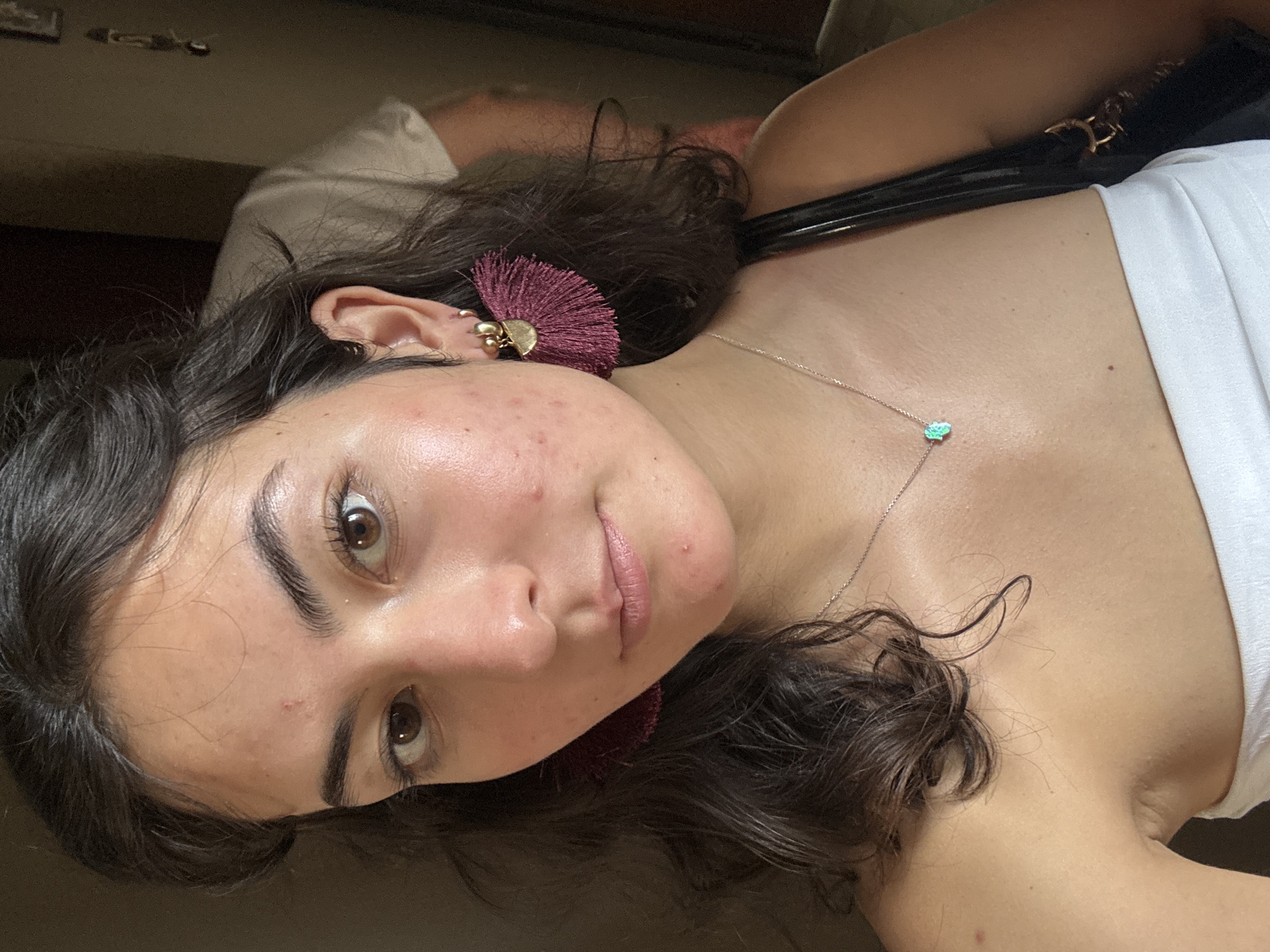 A close-up, rotated selfie depicts a young woman with olive skin, dark brown curly hair, and dark brown eyes. She wears a white top, and a silver necklace with a light blue pendant. Her left ear features multiple piercings and a large burgundy tassel earring with a gold semi-circle base. Her face shows some acne. The background is blurry and appears to be an indoor setting, possibly a home. A glimpse of another person wearing a light-colored garment is visible in the background. The scene does not provide any clear indication of being in Puebla, Mexico.