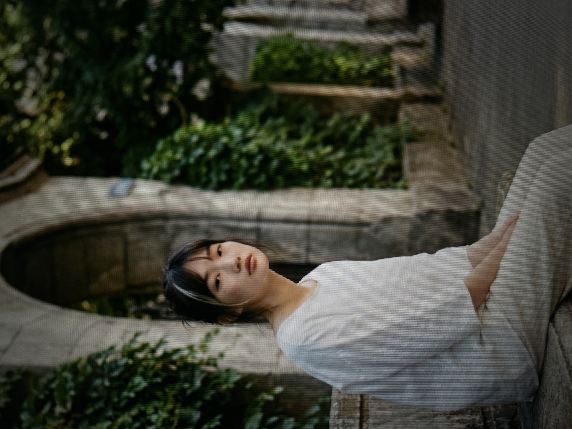 A young East Asian woman with dark hair, styled with blonde streaks and bangs, is sitting on a stone ledge in front of a stone archway covered in ivy. She is wearing a loose-fitting white linen top and matching light-colored pants. Her gaze is directed towards the viewer with a neutral expression. The background features lush green foliage and architectural elements, suggesting an outdoor urban setting with historical features. The lighting is soft, casting subtle shadows.
