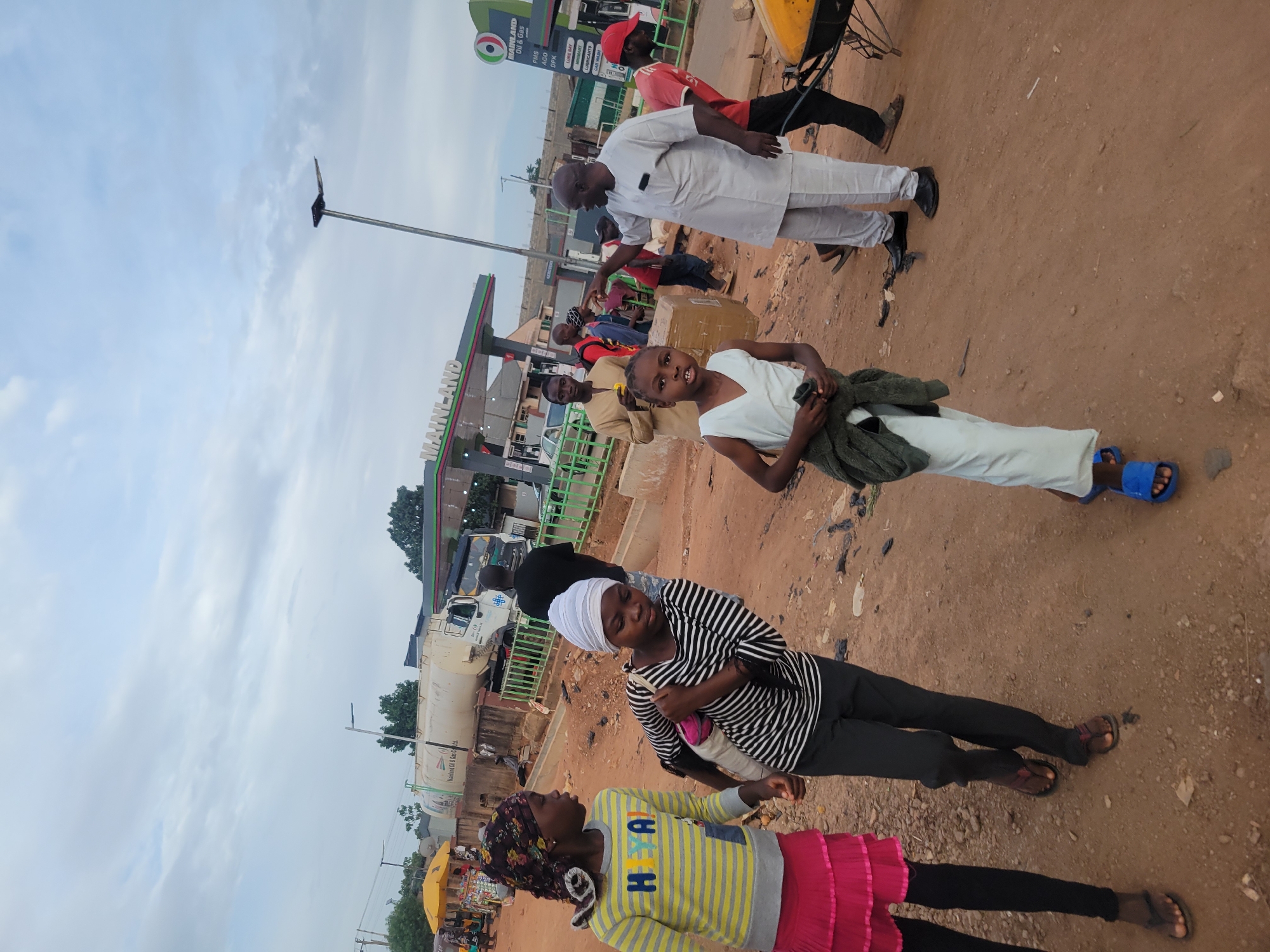 This image captures an outdoor street scene in Jos, Nigeria, depicting several individuals near a gas station under an overcast sky. The ground is a reddish-brown dirt, characteristic of the region.
In the foreground, three females are visible. On the left, a young girl wears a yellow and grey striped long-sleeve top, a pink ruffled skirt, and black leggings with a patterned headscarf. She is looking towards another female in a black and white striped top, black pants, and a white headscarf, who is holding a light-colored bag. Further right and slightly behind them, a young girl in a white top and white pants holds a dark green garment and wears blue sandals, looking directly at the camera.
In the middle ground, a man in a white traditional garment (agbada-like) and black shoes stands with his back partially turned. Behind him, another man in a red shirt and red cap is positioned near a yellow structure, possibly a vendor cart. Several other people are discernible further back near the gas station pumps.
The prominent structure is a "MAINLAND Oil & Gas" station, identified by its signage which reads "MAINLAND Oil & Gas" and lists services such as "PMS", "DPK", "LUBE BAY", "LUBRICANTS", and "CAR WASH". A large white tanker truck is parked near the station. A tall utility pole stands on the left side of the scene.
The cloudy sky suggests soft, diffused light, indicating either an early morning, late afternoon, or generally overcast weather. The scene portrays a casual daily street environment with people going about their activities.