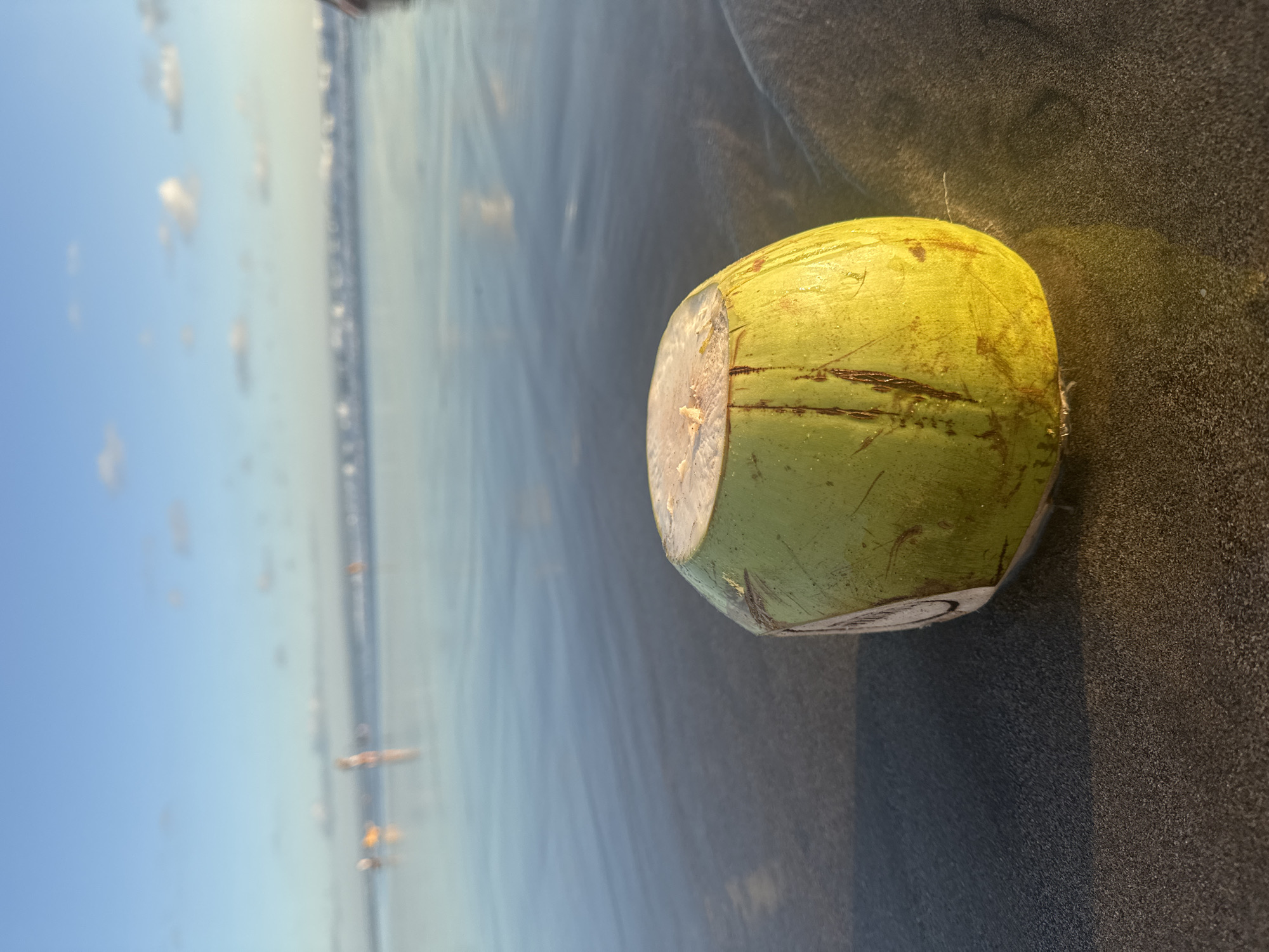 The image depicts a cut coconut on a dark sand beach in North Kuta, Indonesia. The coconut, predominantly green with brown markings, is positioned towards the right side of the frame. The cut top exposes the white flesh. To the left, the scene opens up to a blurred ocean landscape, with people visible in the distance along the shoreline. The sky is blue with scattered clouds. The dark sand is wet, reflecting the sky and ocean.