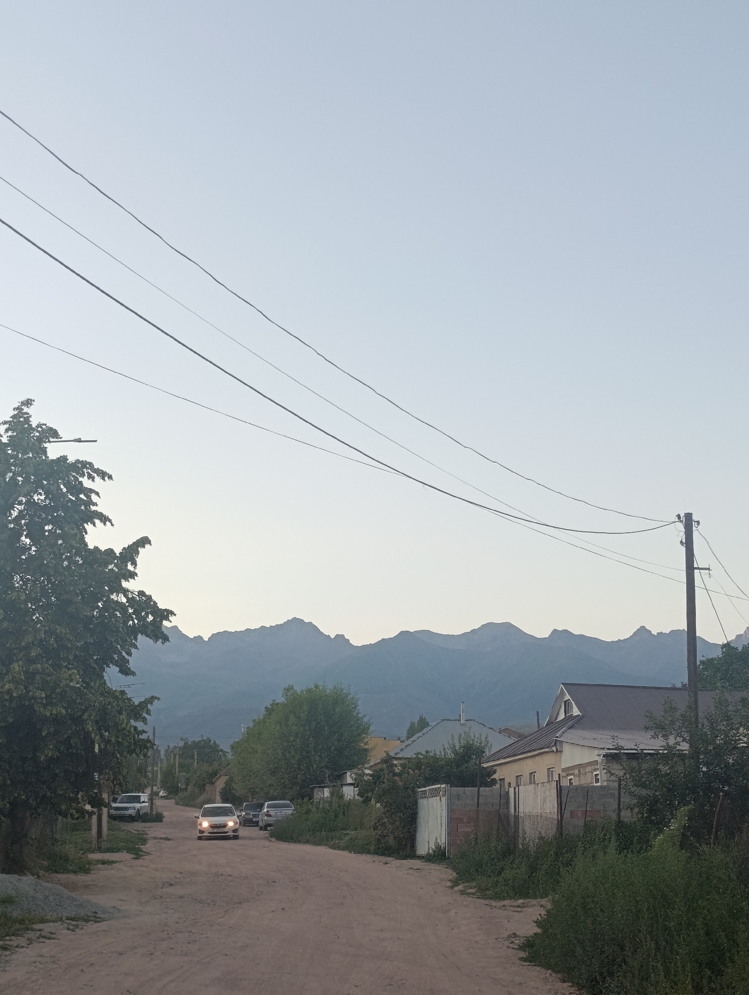 The image depicts a street scene in the city of Bosteri, Kyrgyzstan. A dirt road leads into the frame, lined with houses and trees on either side. In the foreground, power lines stretch across the sky, supported by a wooden pole on the right. Several cars are parked along the road and in driveways, and one white car is driving towards the viewer. In the background, a range of majestic mountains is visible, silhouetted against a clear, pale sky. The overall impression is one of a rural or semi-rural residential area on the outskirts of a city, with the natural beauty of the mountains serving as a striking backdrop.