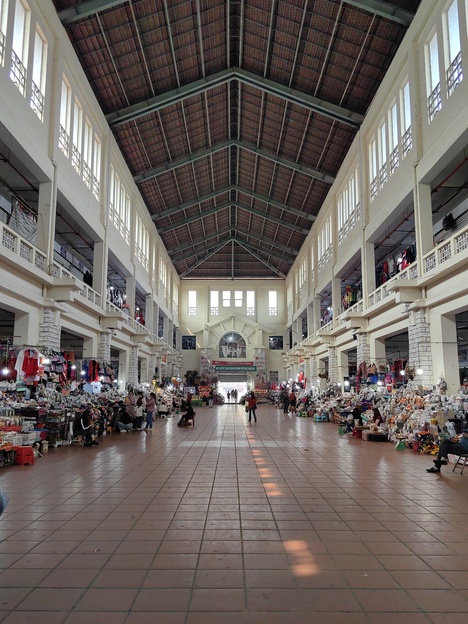 The image depicts the interior of the Sa Pa Market (Chợ Sa Pa) in Sa Pa, Vietnam. The perspective is from a low angle at the front of a wide central aisle, looking towards a prominent arched entrance at the far end.
The market hall is a large, multi-story structure characterized by light-colored, possibly cream or beige, walls and columns. A high, gabled roof with exposed brown corrugated panels and metal trussing is visible overhead. The upper levels on both sides feature numerous tall, narrow rectangular windows with simple decorative metal grilles, allowing natural light into the space. Balconies with decorative railings run along the upper levels, from which clothing and other merchandise are draped. Red pipes are visible beneath the ceilings of the upper levels.
The ground floor on both sides of the central aisle is densely occupied by vendor stalls. These stalls display a vast array of goods including clothing, bags, shoes, packaged items, household articles, and souvenirs, piled on tables, hung from racks, and arranged on the floor. The stalls utilize rudimentary setups with tarps, crates, and small plastic stools.
Approximately twenty individuals are visible throughout the market. In the mid-ground, a cluster of people, including adults and children, is gathered around a stall on the left, potentially browsing merchandise. Another individual is seated on a chair on the right. Towards the far end, several figures are walking towards or through the main arched entrance. A person in a red jacket walks away from the camera in the mid-left. Most people are engaged in shopping or vending activities.
The central aisle features a floor of brown square tiles, with diagonal patches of sunlight reflecting off the surface in the foreground. A large banner with Vietnamese text, including "CHỢ SA PA" (Sa Pa Market), and imagery hangs above the arched entrance at the rear, flanked by additional rectangular windows. The scene is well-lit by a combination of natural light from the high windows and localized artificial lighting at the stalls.