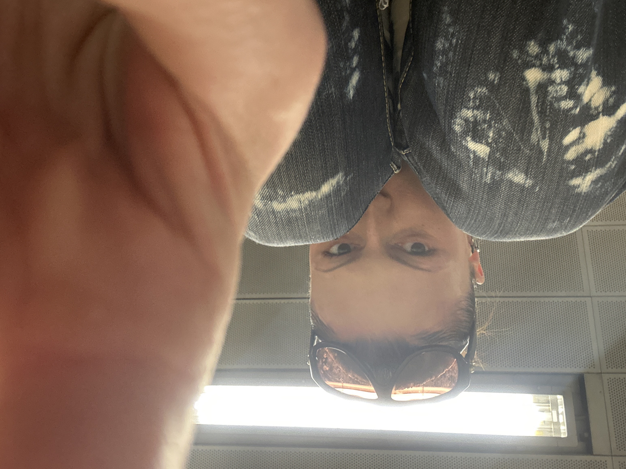 A close-up photograph, presented sideways, depicts an individual in what appears to be a modern public transport or indoor terminal environment within the City of London, United Kingdom. A large, out-of-focus human hand occupies the top and upper-right section of the frame, suggesting a close-proximity shot. The subject, positioned centrally, has their head tilted towards the right side of the image, with dark hair pulled back. They wear dark-framed sunglasses with reddish-brown tinted lenses pushed up onto their head. Their eyes are open and directed towards the camera. The individual is dressed in dark blue denim jeans featuring pronounced bleached or acid-wash patterns. The visible background consists of light grey, uniformly perforated paneling. On the left, a prominent vertical rectangular light source, possibly a window or an illuminated fixture, emits intense white light, obscuring details beyond it, but suggesting an exterior view or an adjacent space.