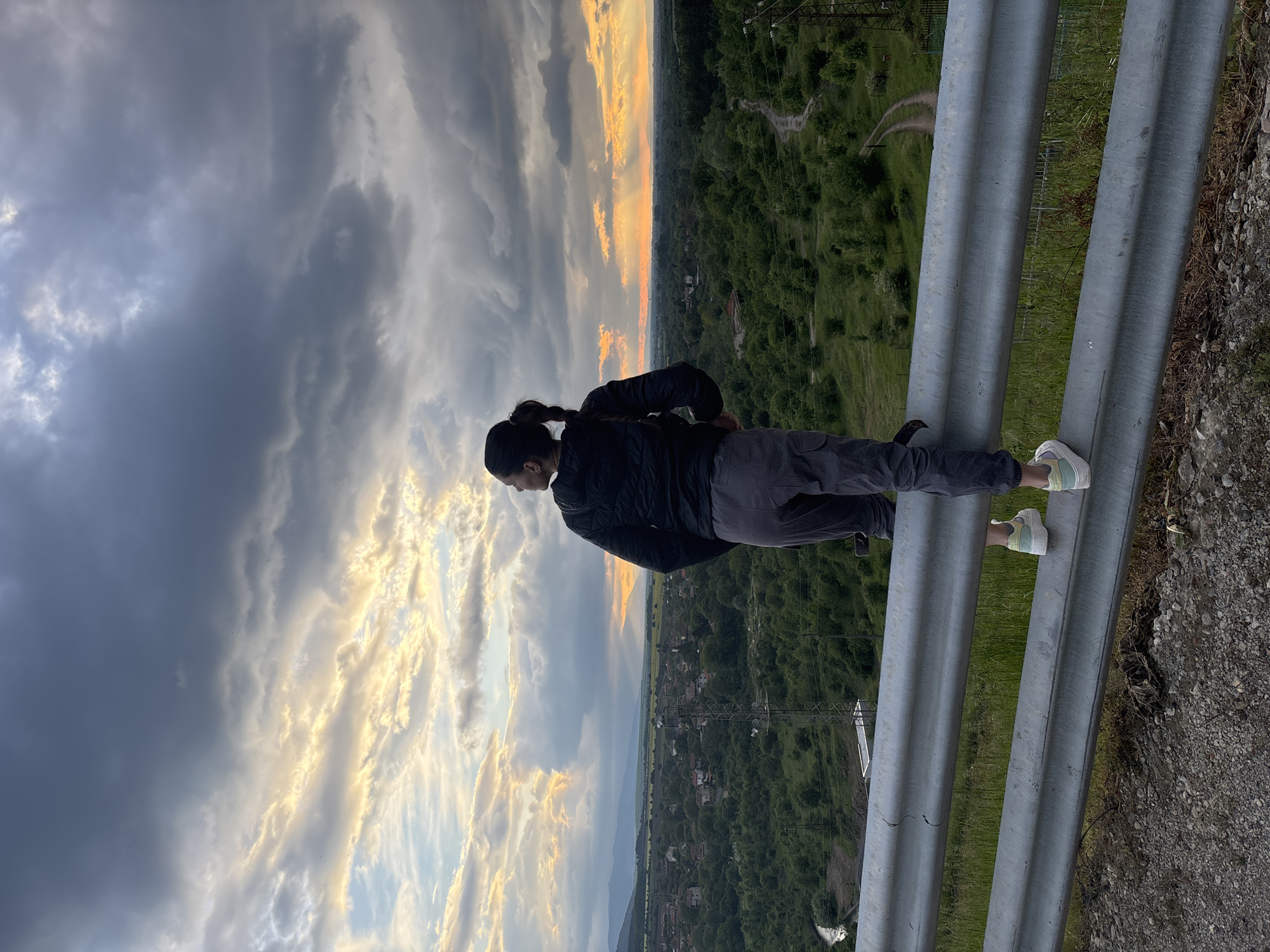 A landscape-oriented photo depicts a woman standing on a metal guardrail overlooking a valley at what appears to be dusk or dawn, taken in or around Ognyanovo, Bulgaria. She has dark hair pulled back in a ponytail and is wearing a dark jacket and gray pants with light-colored shoes. She is facing the valley. The foreground consists of the gray metal guardrail, and the background features a broad valley with trees, buildings, and a dramatic sky with contrasting dark and light clouds and a strip of orange and yellow horizon light. A cellular phone is tucked in her left pocket.