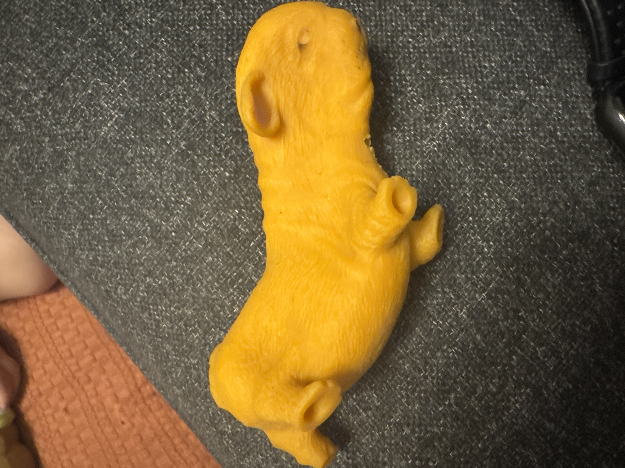 An orange, plastic toy animal resembling a guinea pig lies on a dark, textured surface. The toy is lying on its side with its head facing to the right. It has two front paws and two hind legs, all of which appear bent. The toy has two small ears on its head and what appear to be two small eyes. The surface it is on is dark gray with a flecked texture. In the upper left corner of the image, a small portion of an orange, woven material is visible, along with a small section of a person's leg.