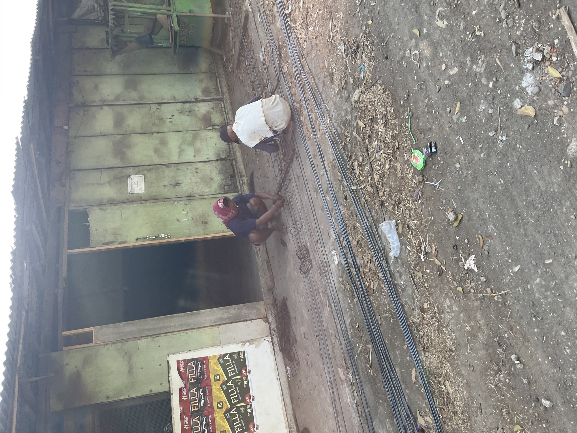 In Pelang, Indonesia, a rough, outdoor construction site is depicted. Two individuals are present, actively engaged in manual labor. One person, wearing a red headscarf and a dark shirt, squats on the ground, seemingly working with metal rods. Another individual, dressed in a dark shirt and white garment, also squats, focusing on a mesh of metal bars. The environment is characterized by a weathered, green-painted building with a corrugated metal roof. Several long metal rods are laid out on the ground, indicating ongoing construction work. Debris, including dry leaves and plastic, is scattered across the dusty ground. A poster advertising "Filla SPR" is visible on the building's exterior. The scene suggests a basic, possibly informal, construction or repair project in a community setting.