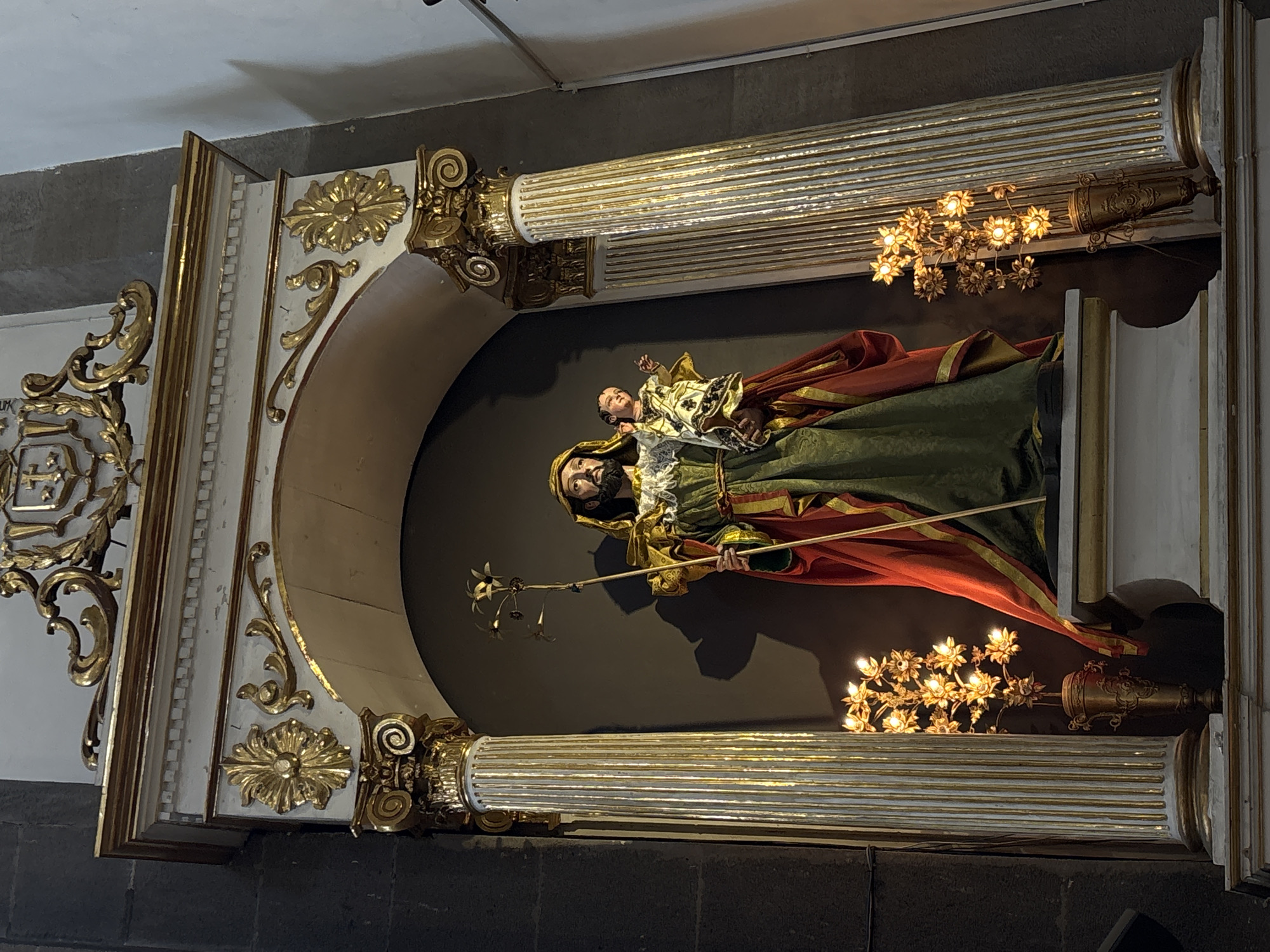 The image captures a statue of Saint Joseph holding the child Jesus in the city of Puebla, Mexico. The statue is positioned within an ornate, gilded alcove with Corinthian-style columns. The statue itself is adorned with robes of green and red, and Joseph is depicted holding a staff with lilies. Floral chandeliers flank the statue within the alcove. The alcove is set against a dark background. The photograph is taken indoors, likely within a church or religious building.