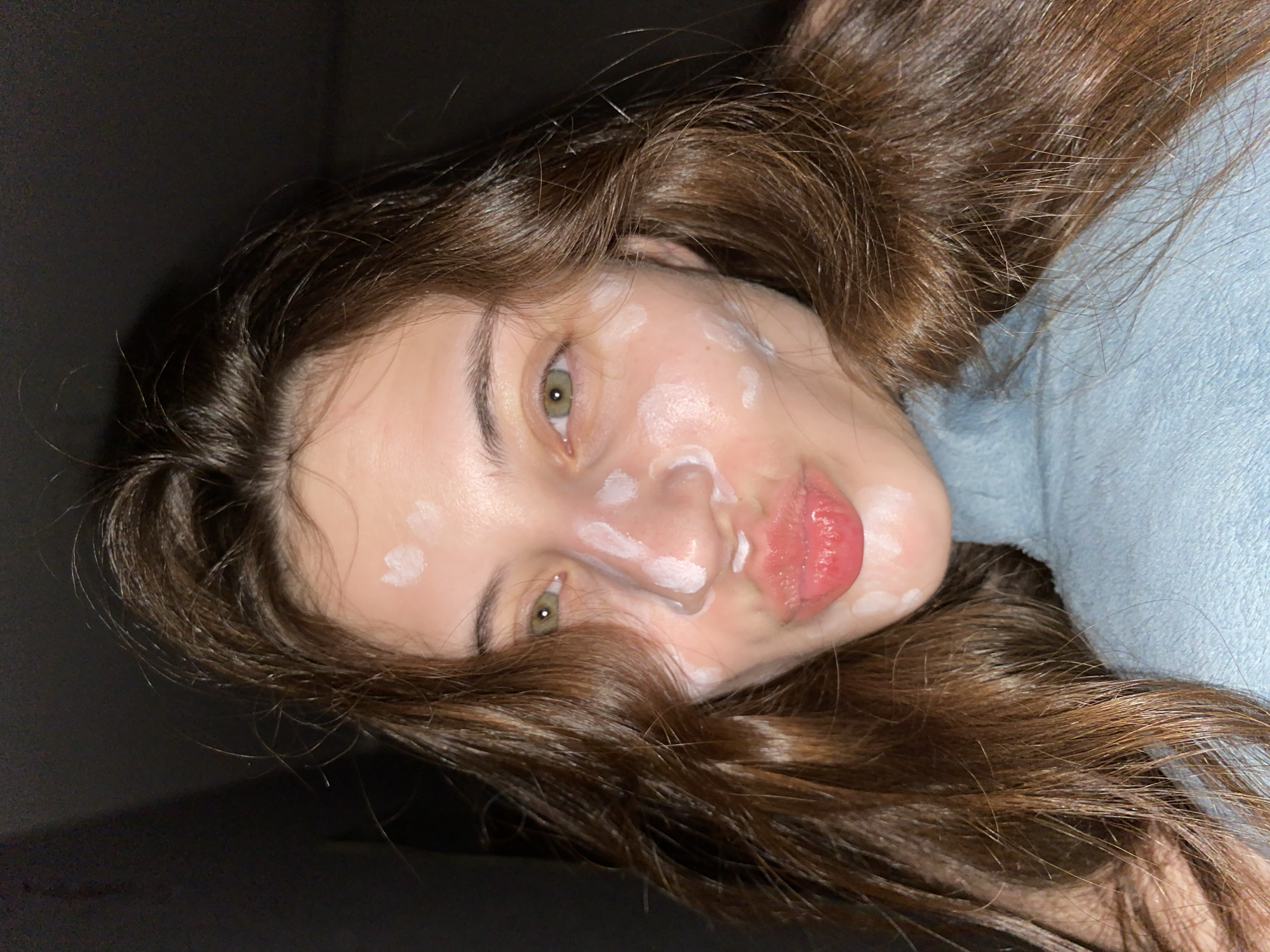 A young woman with long brown hair and green eyes lies at an angle in a dimly lit room. She has white cream applied in spots on her face, primarily around her nose and cheeks. She is wearing a blue garment. The location cannot be determined to be Votkinsk, Russia, based on the visual information provided.