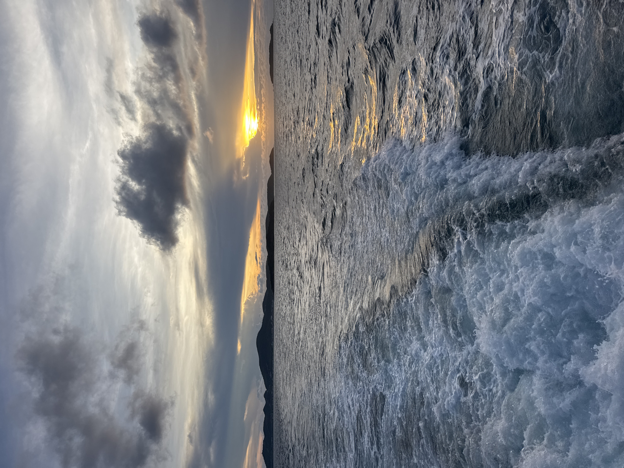 The image shows a body of water, possibly the sea, with choppy waves and a foamy wake on the right side of the frame. In the background, there is a sunset with the sun obscured by clouds, casting a golden glow across the sky and water. A dark, mountainous or hilly landmass is visible on the horizon. It is likely that the location is Pattaya City, Thailand, given the coastal environment and the presence of a sunset over water. The image appears to have been taken from a moving vessel, as evidenced by the wake in the water.